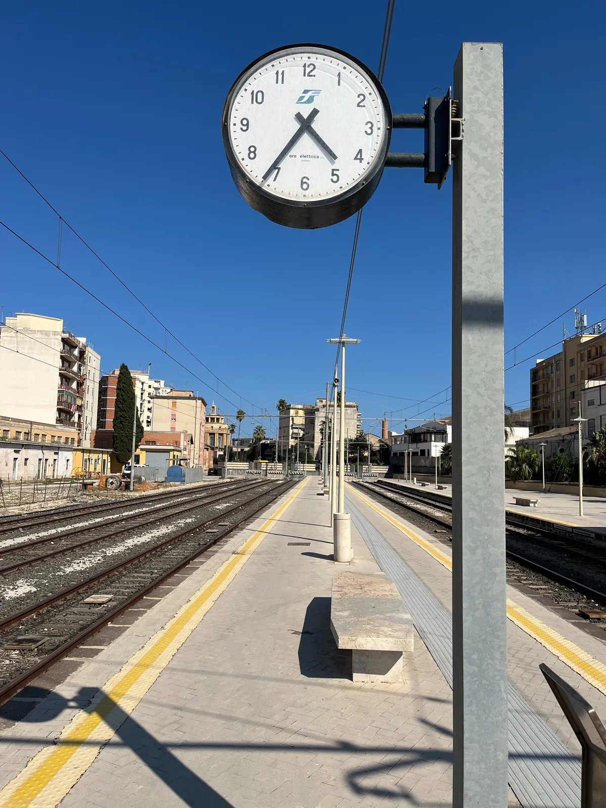 Standing on an empty platform between the train tracks. In the background there are some appartment buildings. The clock on the platform points at 24 minutes to 5.