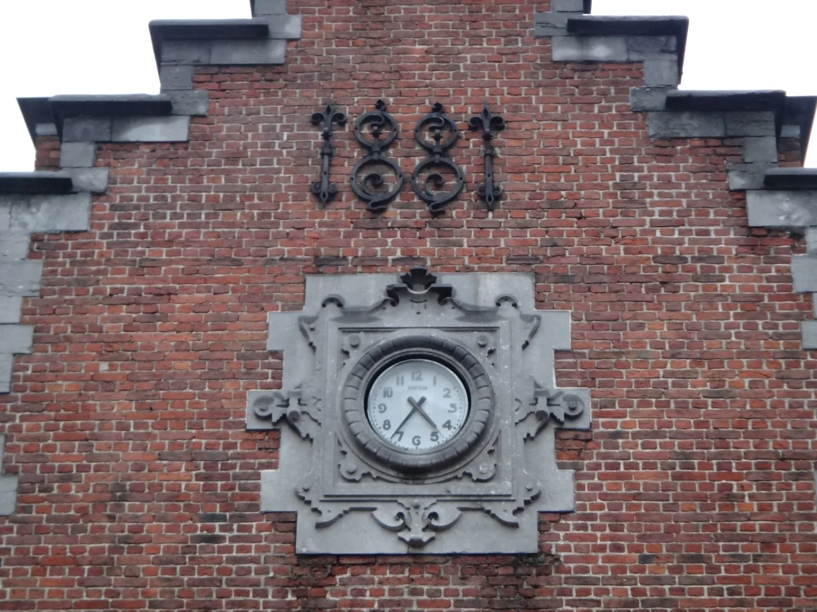 In the facade of a step-roofed building, which, according to the cast iron letters was built in 1881, there’s clock. It points at 36 minutes past 4.