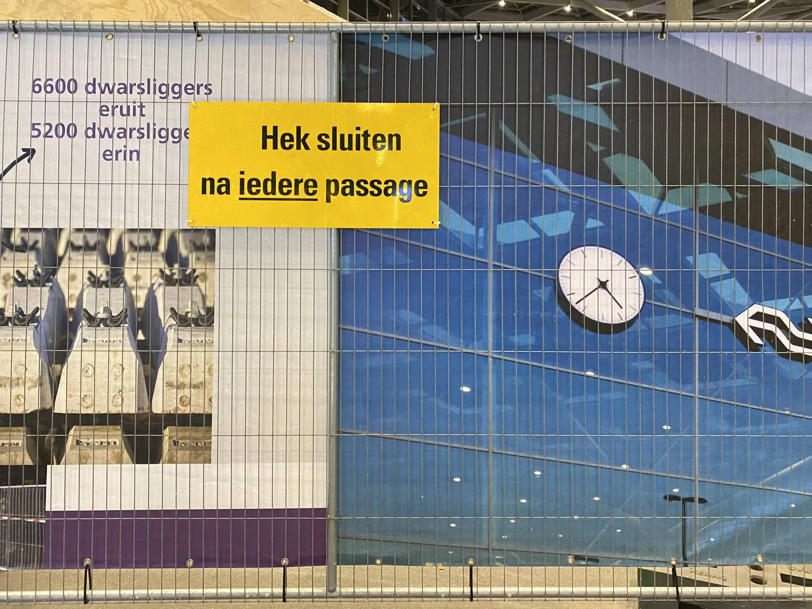 There are large photographs behind this fence. There’s a sign on the fence that says, in Dutch, close the fence after every passage. On one of the large photographs there’s a clock which points at 23 minutes to 5.