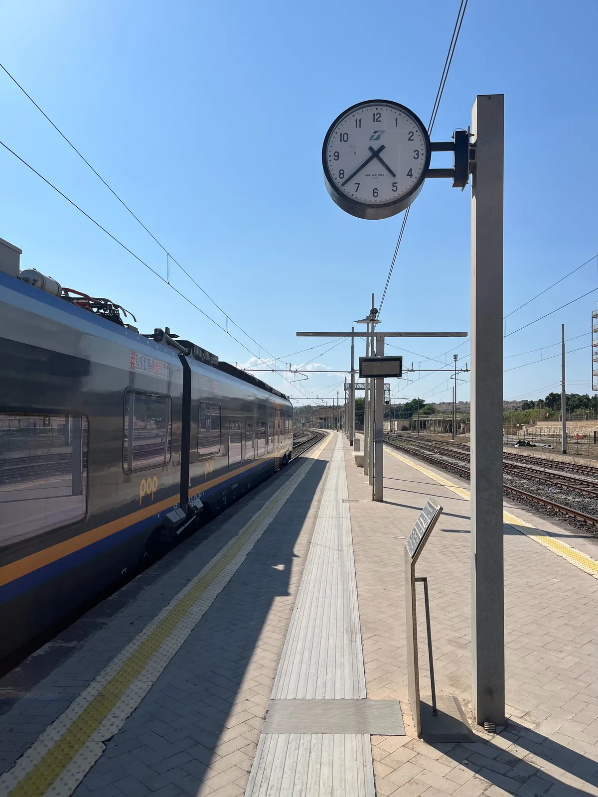 A bright blue, summery sky. On the left there’s a train standing still on the platform. On the right there’s a deserted landscape. In the middle there’s a clock that points at 34 minutes past 4.