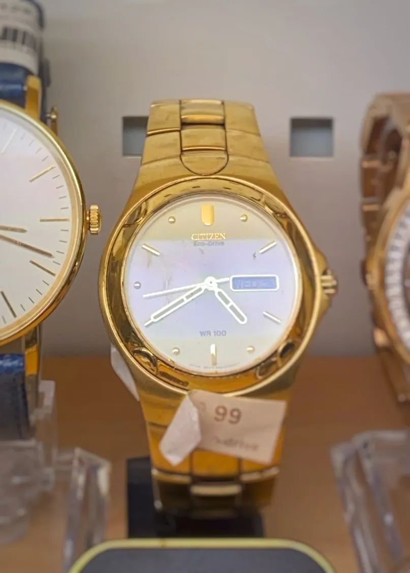A golden wrist watch. It says Citizen Eco Drive WR 100 on its face. It points at 20 minutes to 5.