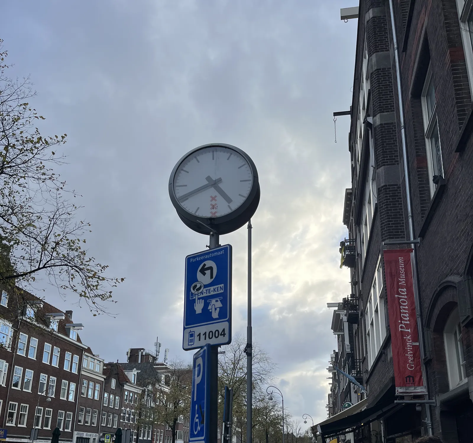 A clock on a pole in a street in Amsterdam, next to the Geelvink Pianola Museum. It’s a cloudy day. The clock points at 19 minutes to five.