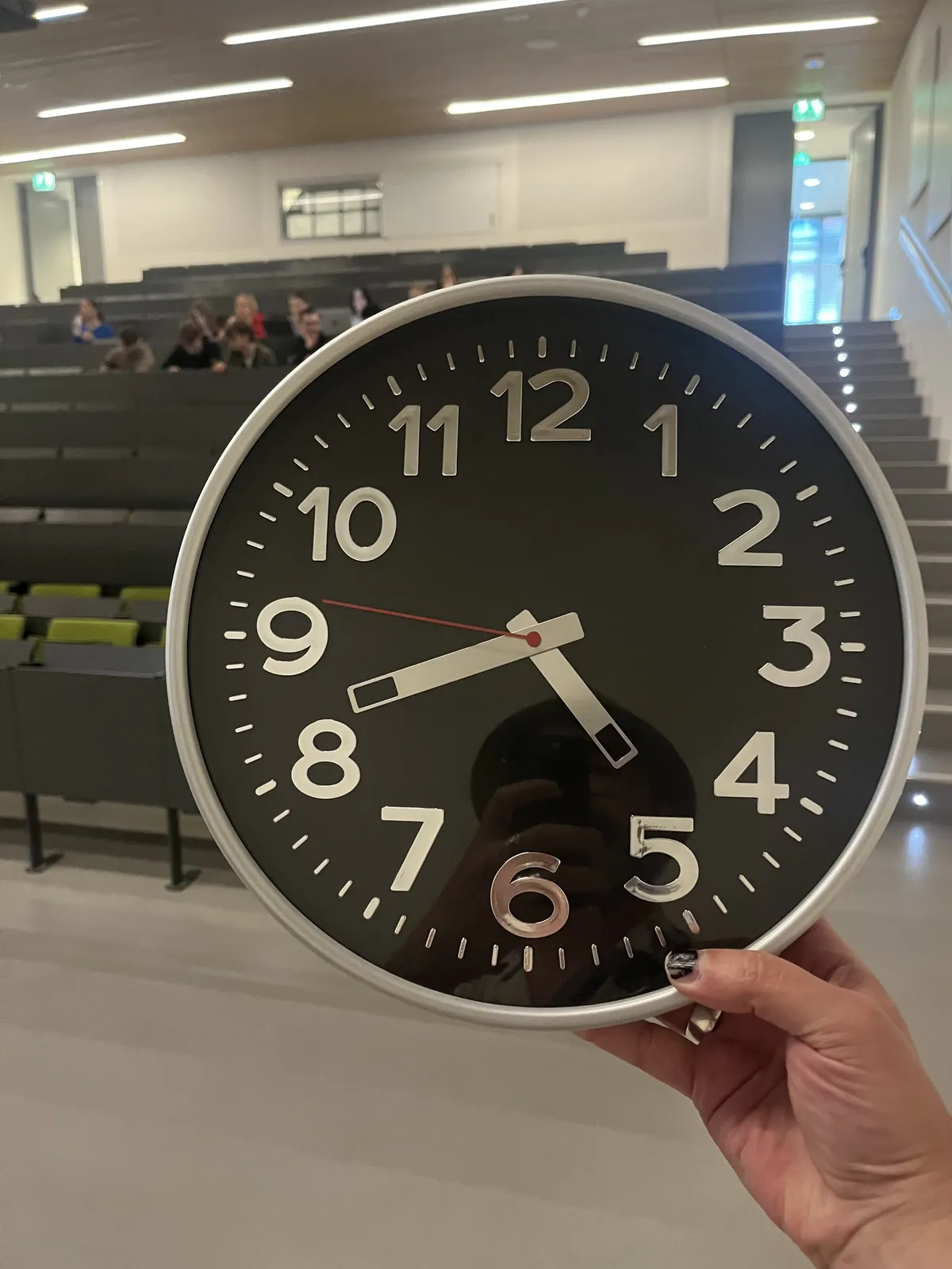 Someone’s holding a black, modern wall clock in their hand. In the background, a bit blurry, a group of people can be seen in a large class room. The clock the person is holding points at 19 minutes to five.