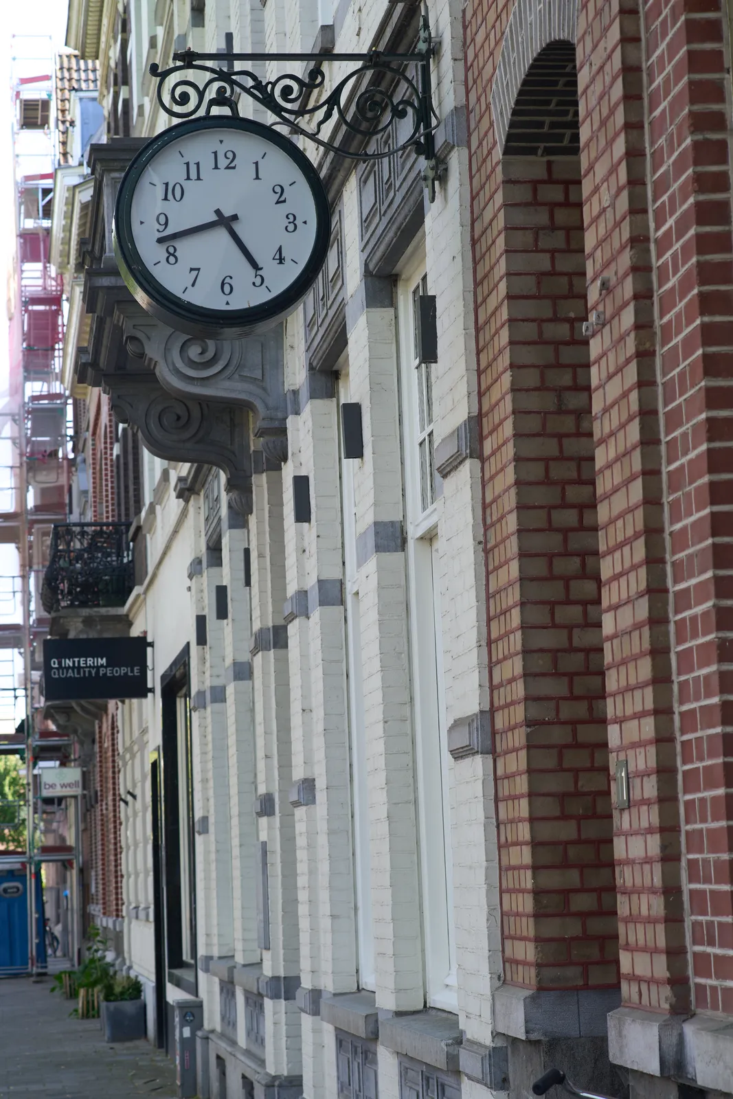 A clock hanging from a cast iron thing, attached to a neo-classical facade. It points at 18 minutes to five.