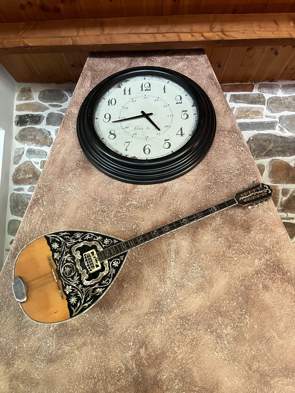 A bouzouki a hanging on a wall, below a large clock. This clock points at 17 minutes to five.