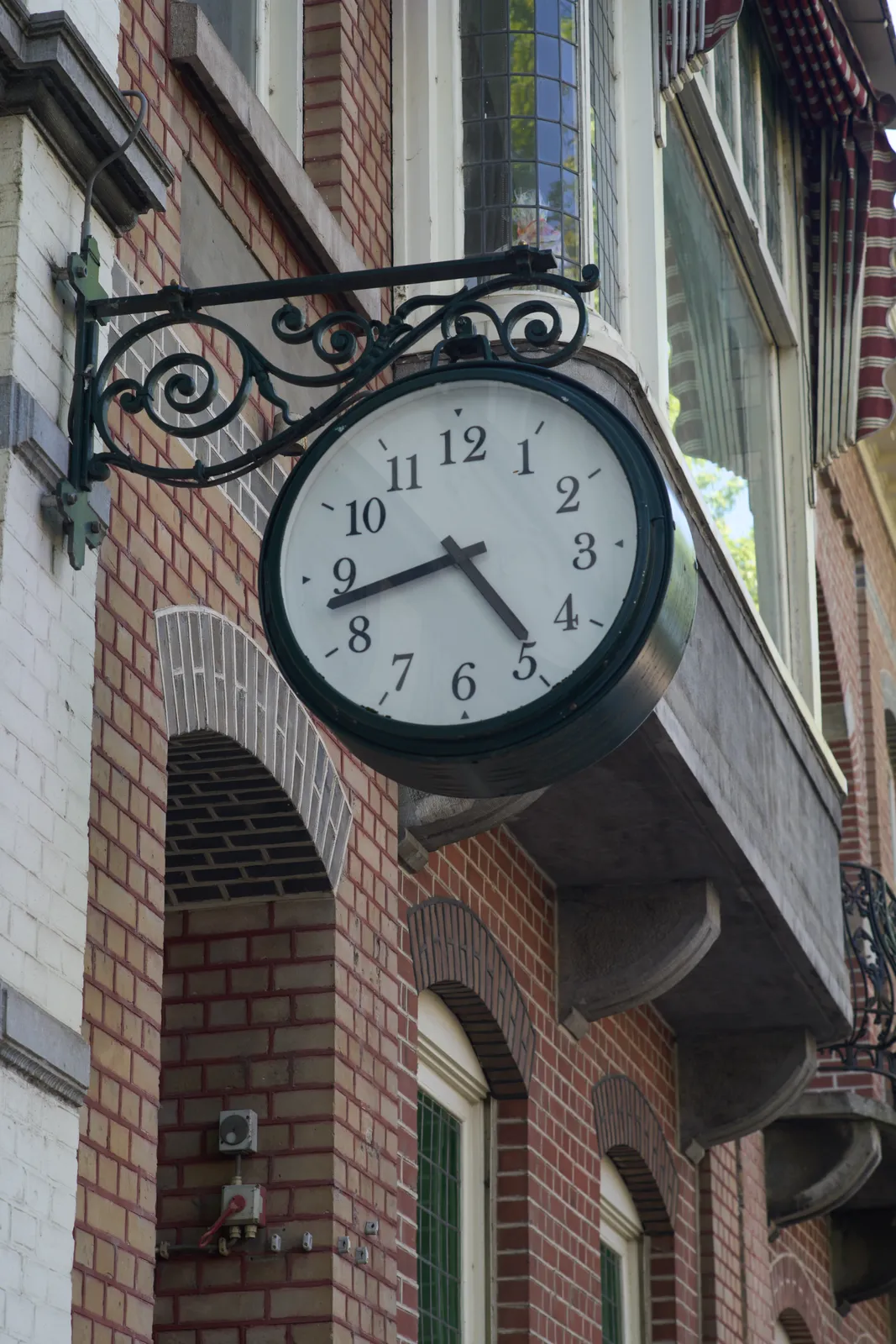 A clock hanging from a cast iron thing, attached to a neo-classical facade. It points at 17 minutes to five.