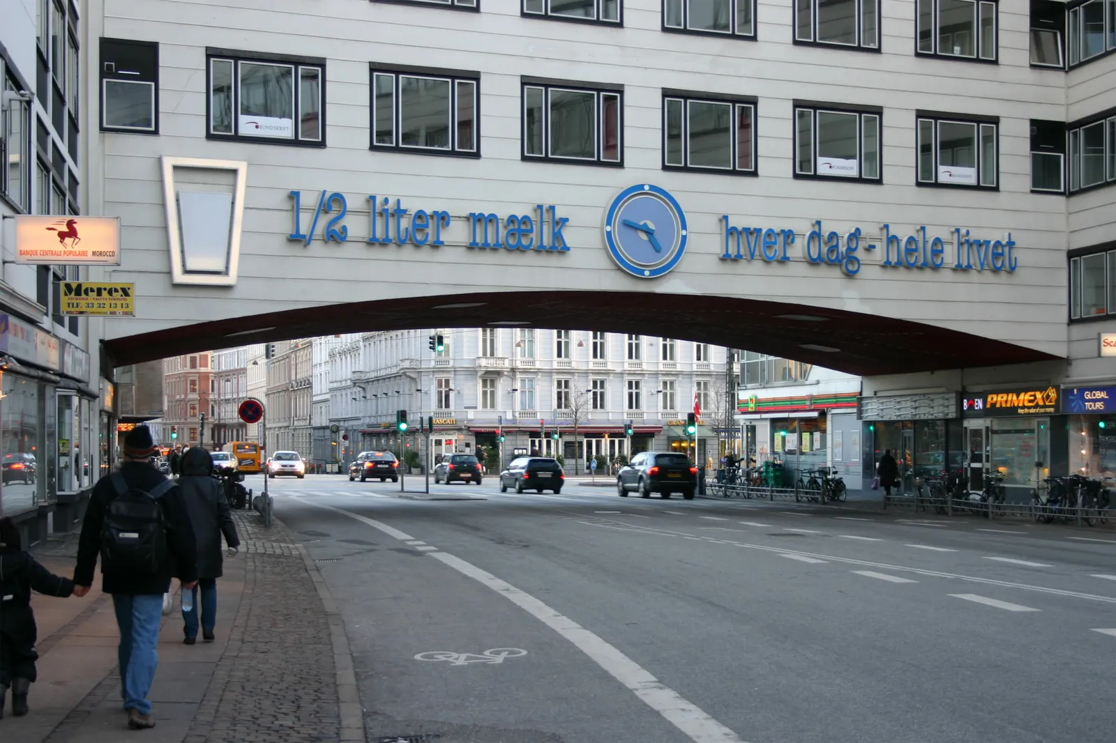 A clock on a building that’s also a bridge over a street. It points at 13 minutes to 5. On the building it also says, in very large type: 1/2 liter maelk hver dag-hele livet