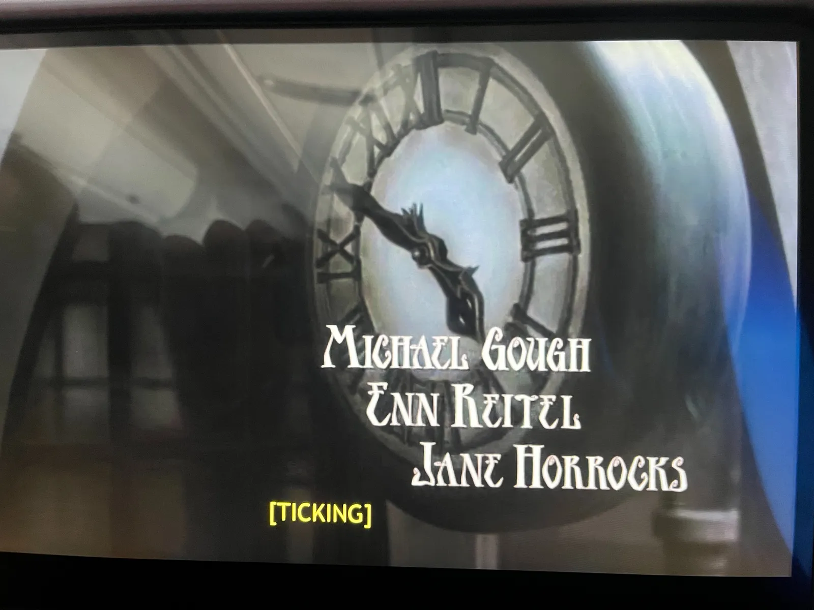 A film still, from the opening sequence of a movie. Three names are on screen: Michael Gough, Enn Reitel, Jane Horrocks. The caption says a single word: ticking. A large clock fills the screens. It points at 10 to 5.