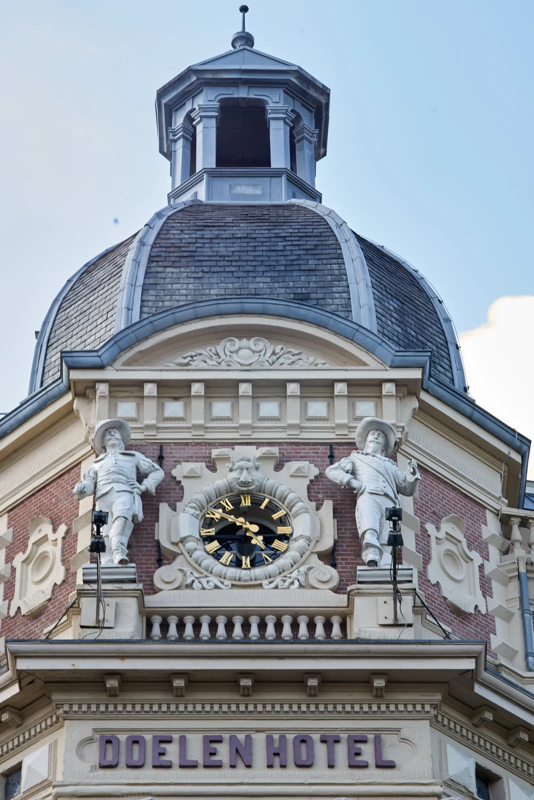 The rightly ornamented tower of the Doelen Hotel in Amsterdam. Between two statues there is a clock. It points at 9 minutes to five.