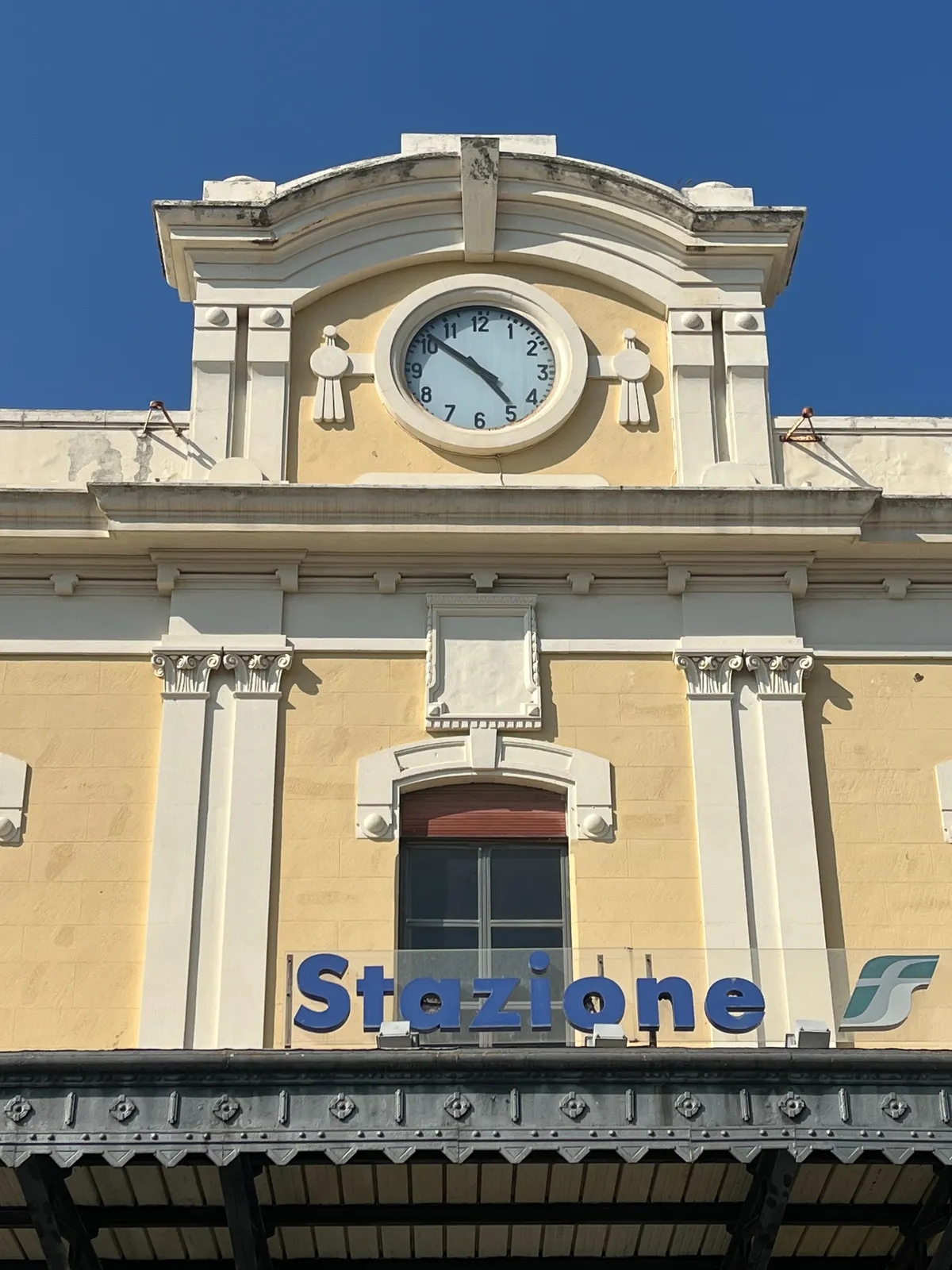 The facade of a train station in Italy (it says Statione in large letters.) There’s a clock that points at 9 minutes to 5.
