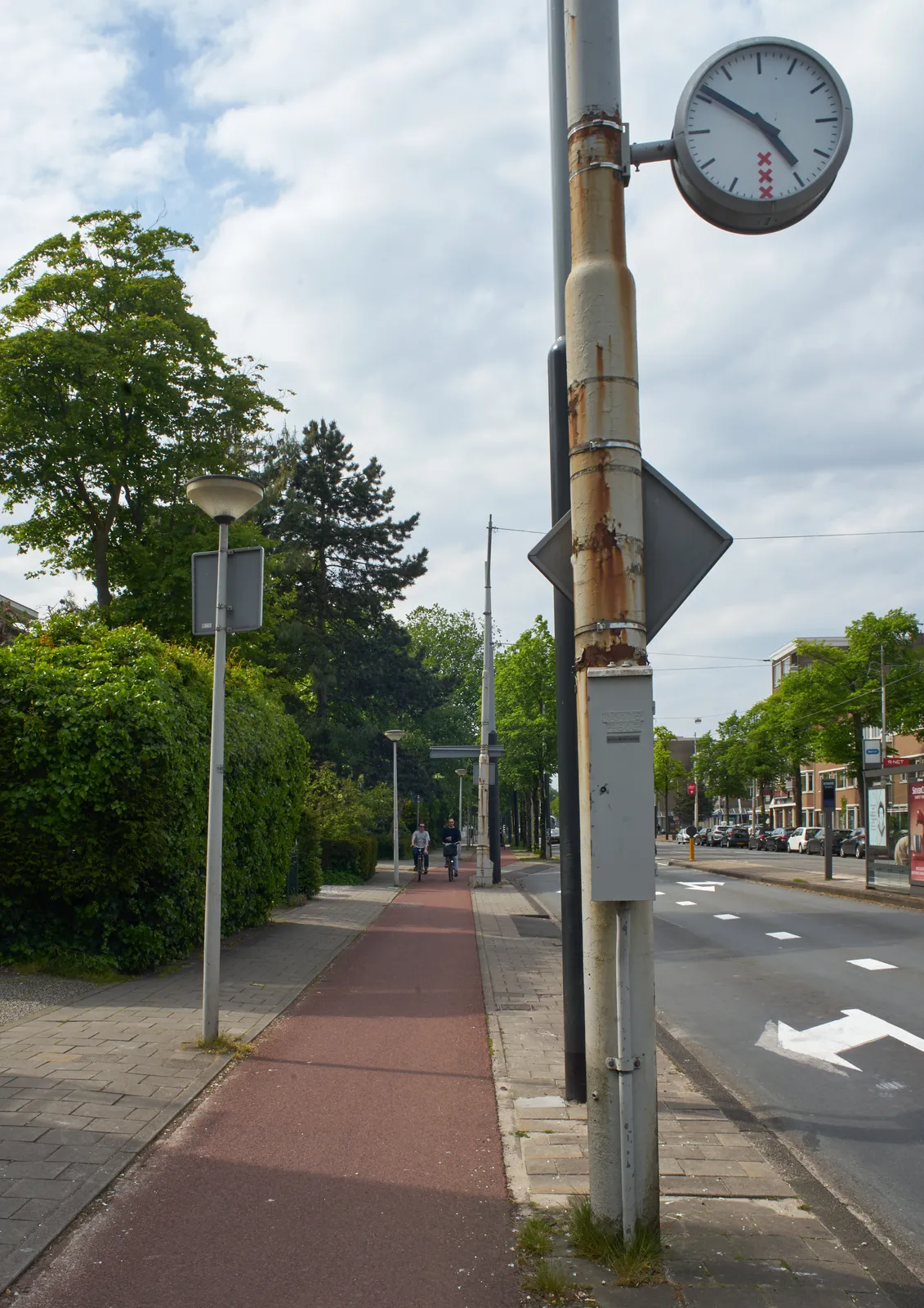 A spring scene in Amsterdam. A car lane on the right, a bike lane — with two people on bicycles — on the left. In the middle there’s a rusty pole with a clock attached to it. It’s 9 to five on this clock.