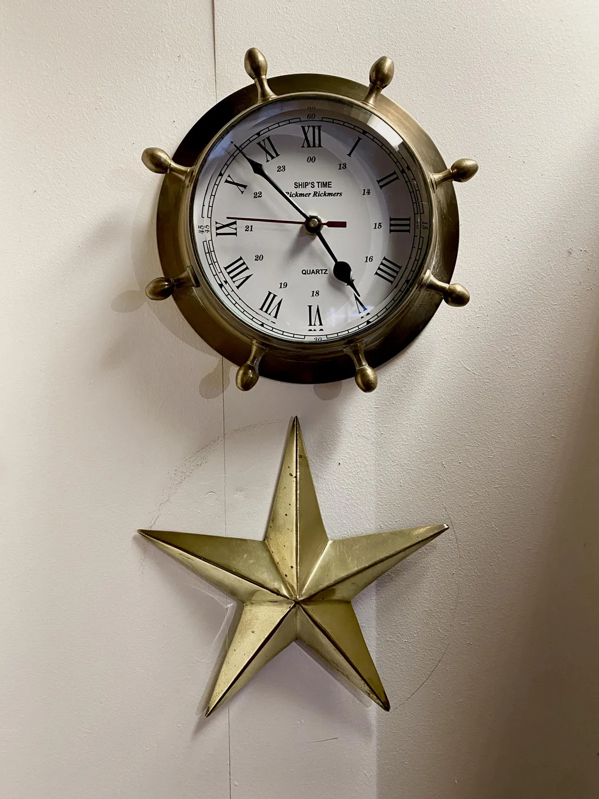 A clock, shaped like the helm of a boat. Below it there’s a star with five points. This star is hanging perfectly centered in a circle. And this circle seems to caused by the clock after it was removed from its previous spot, to be replaced in this new spot a bit higher on the wall. It’s subtle, but it’s visible. The clock points at 8 minutes to 5.