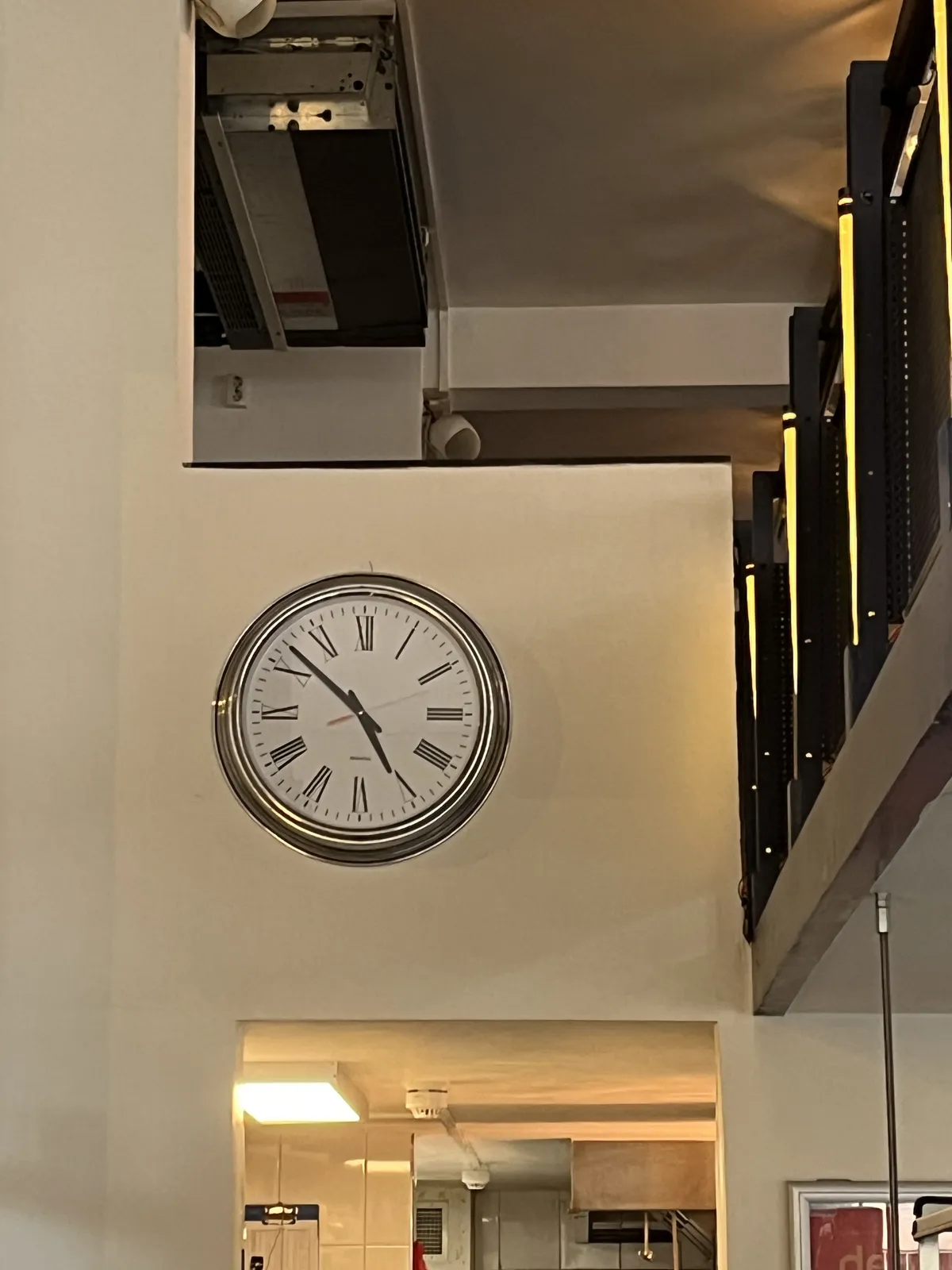 A clock on a wall above a door, and below some sort of balcony, but inside. The clock points at 8 minutes to five.