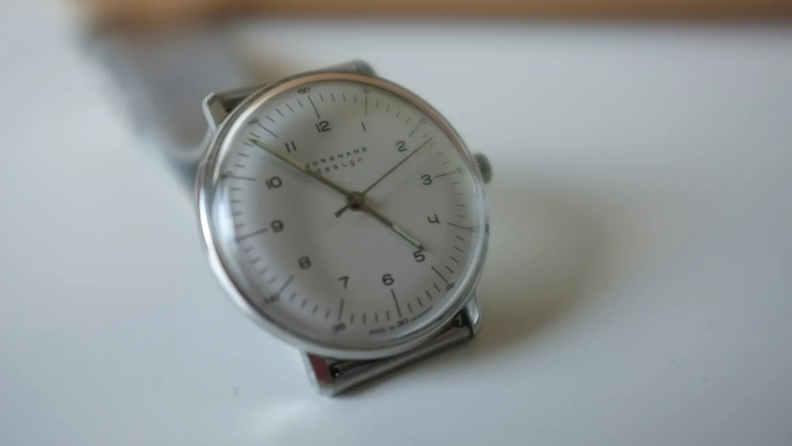A wrist watch, on a table. It says Junghans Design on its face. It points at 7 minutes to 5.