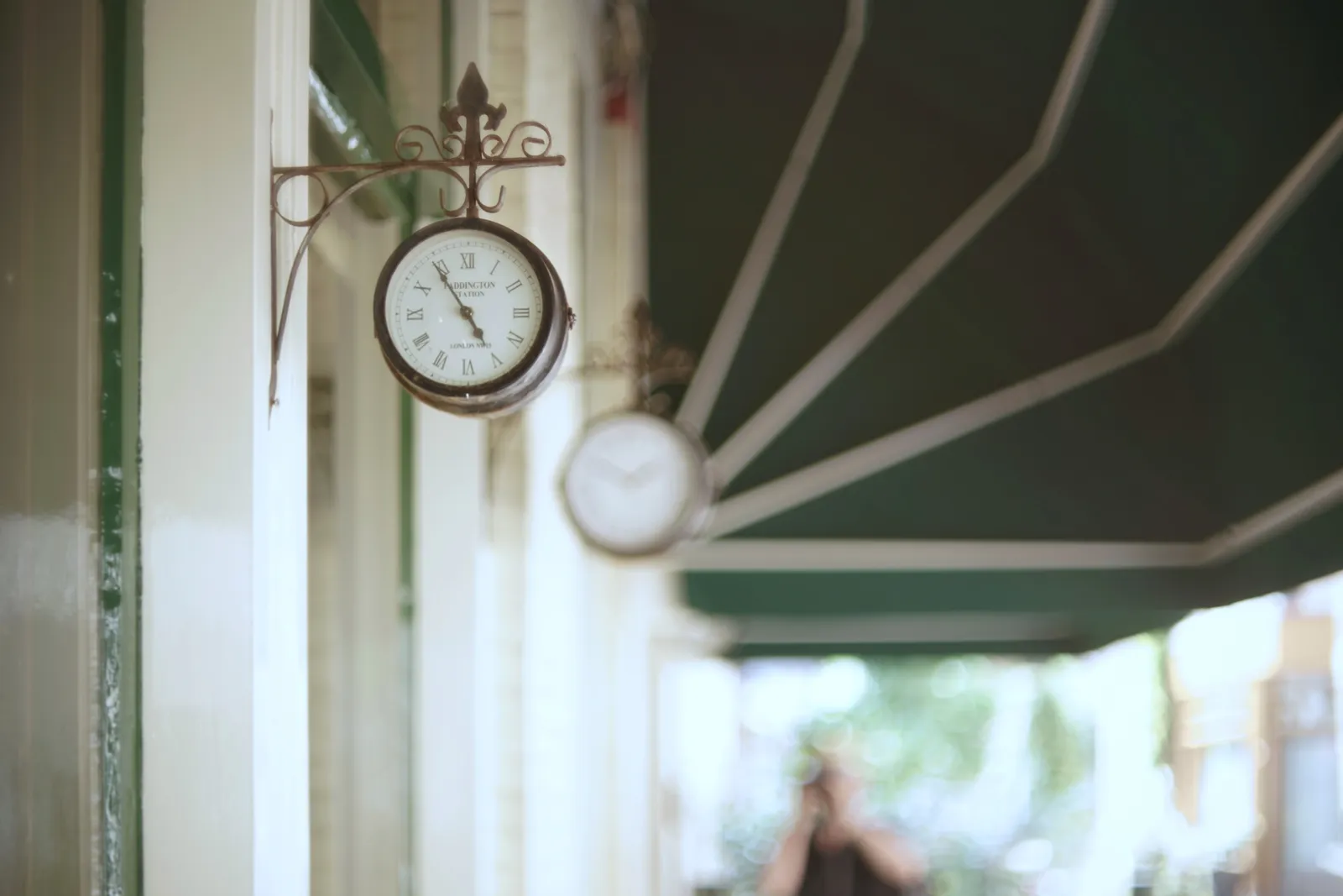 There are two clocks attached to this cafe. The one in front, which is in focus, points at six to five. In the background, very blurry, a photographer can be seen.