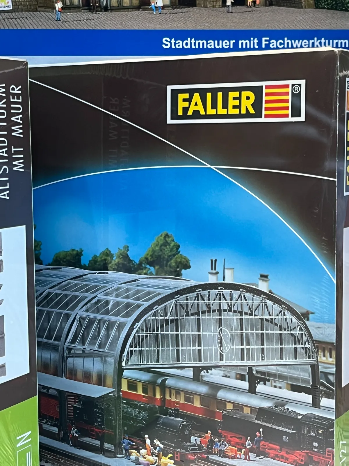 A box with a picture of a miniature version of a train station. On this miniature station there’s a clock which points at 5 minutes to 5.