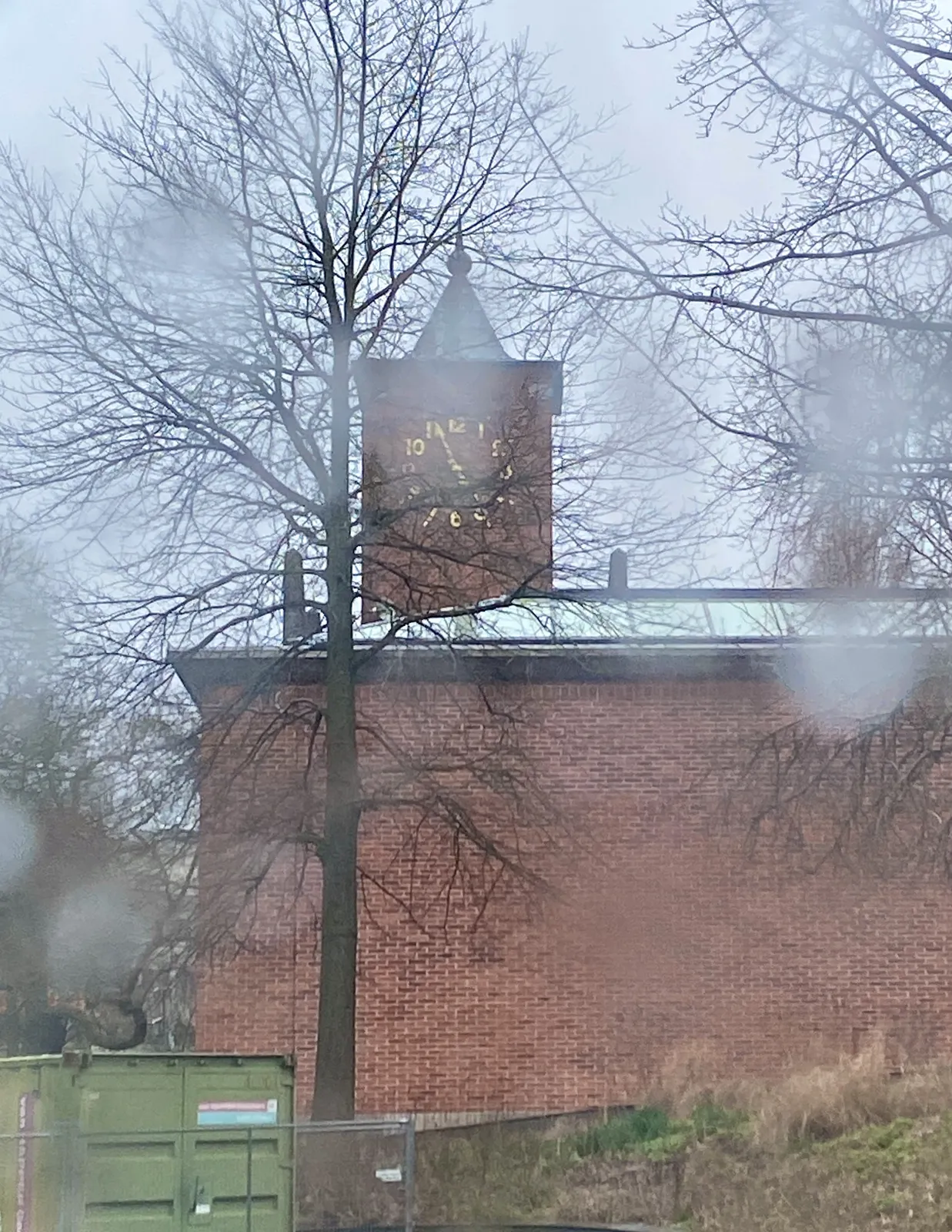 Looking through a window with raindrops on it. Looking at a brick church with a clock. It points at 4 minutes to 5.