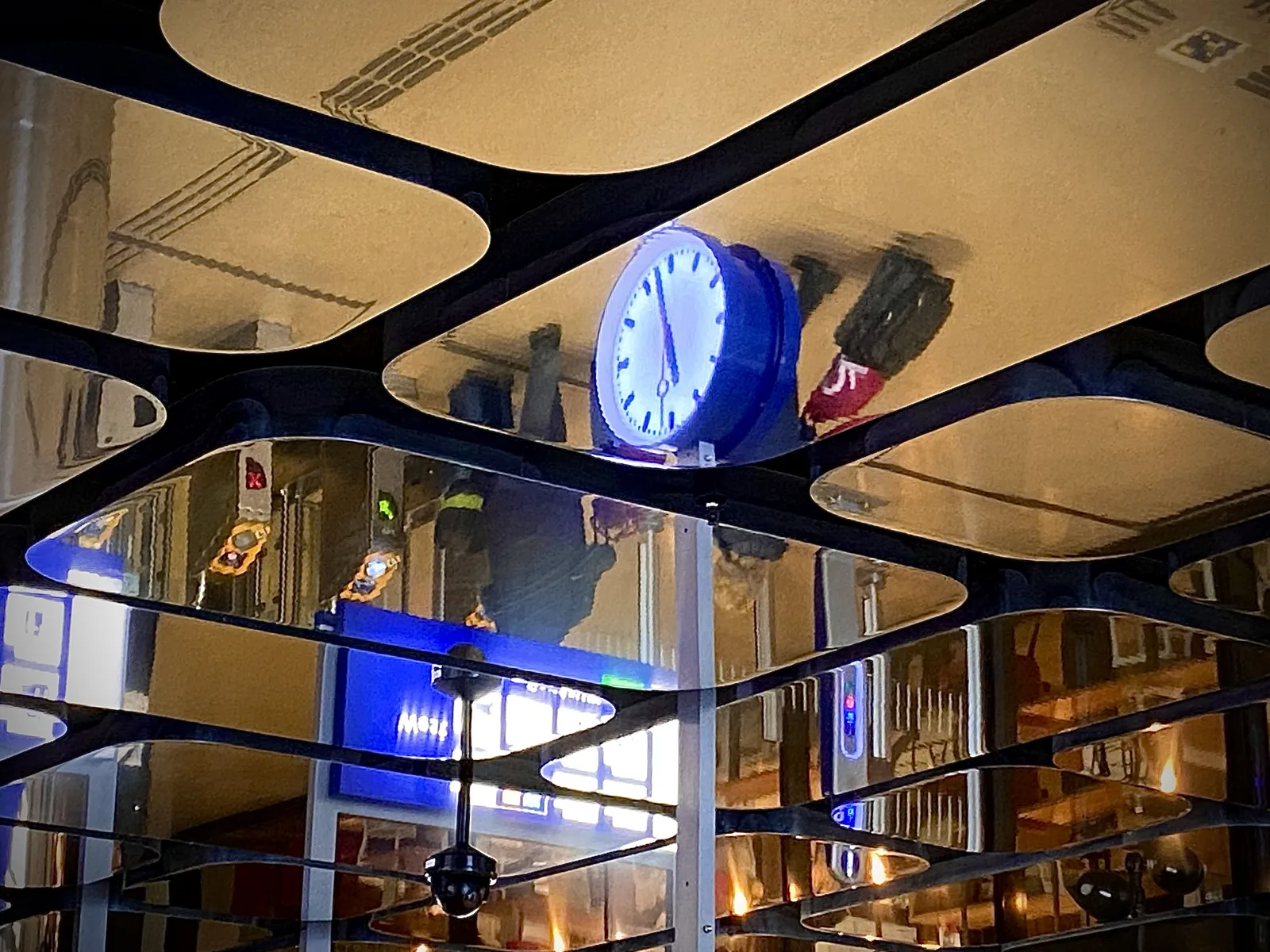 In a mirrored ceiling made of blob-like mirrors, you can see the reflection of a clock in it. It is very difficult to see what time it is exactly, so I won’t even try. It seems to point at approximately 3 minutes to 5.