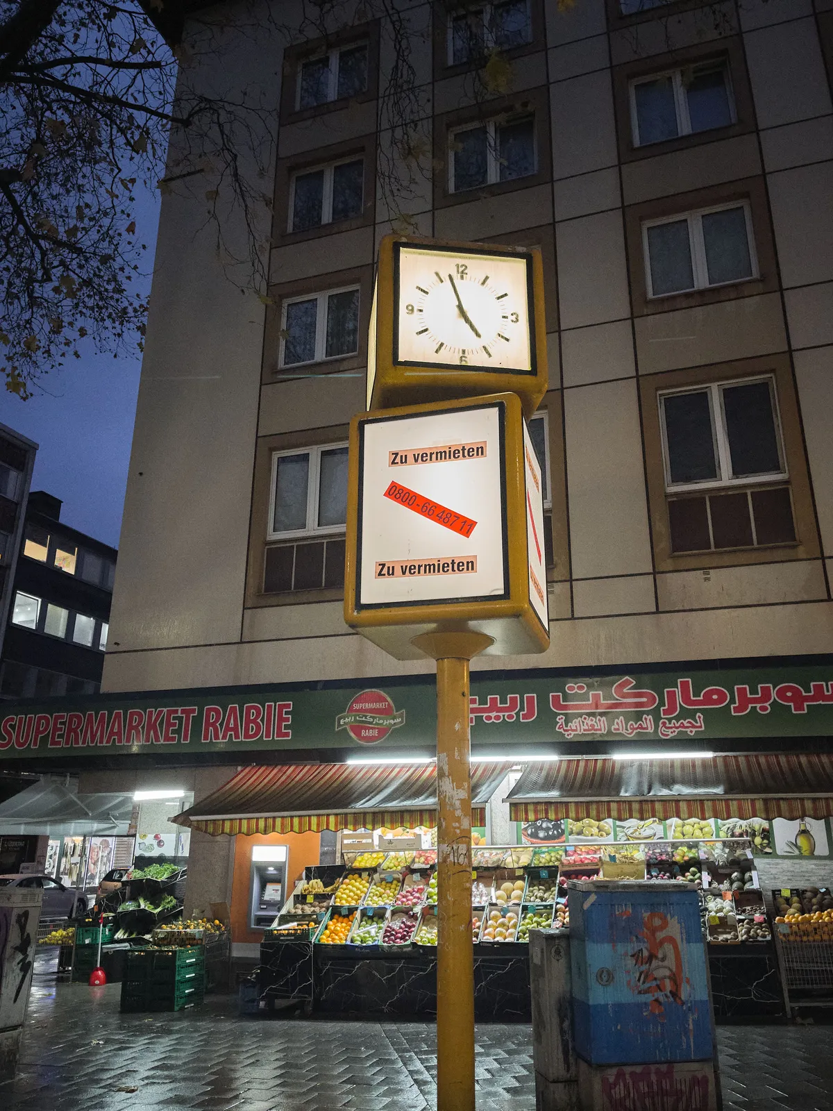 In front of a large, white appartment building, with on the ground floor Supermarket Rabie, stands a pole with a clock, and an empty advertising opportunity, which is for rent. The clock on the pole points at 3 minutes to 5.