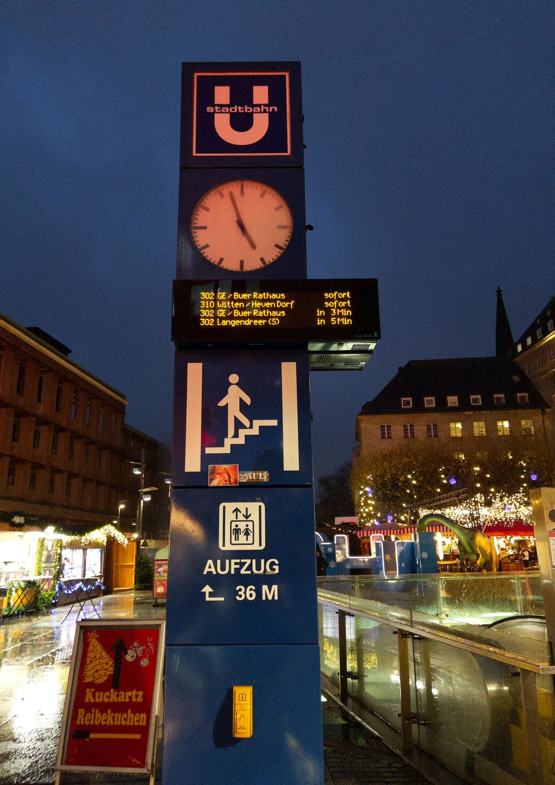In the background you can see a christmas market on a typical German town square. In the foreground a large pole with all kinds of public transport information on it. There’s also a lit clock on this pole, which points at 3 minutes to 5.