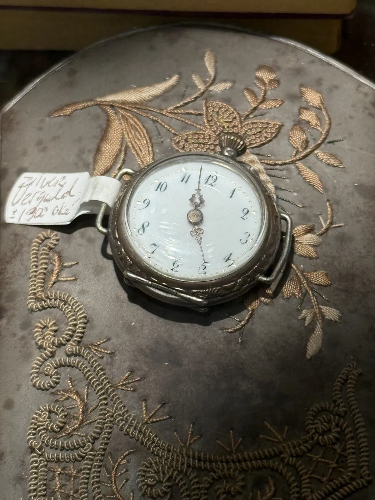 A silver watch, on a gray embroidered rug. It points at three minutes to five.