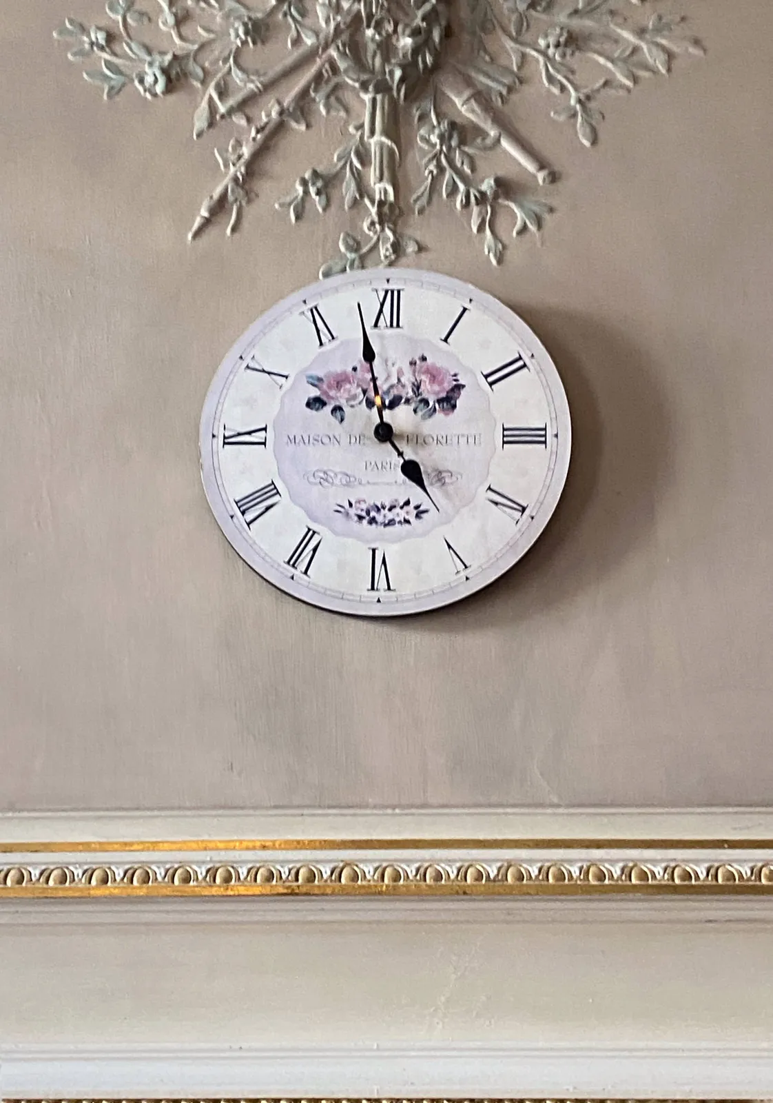 A clock on a decorated wall. There are flowers on its face. It points at 2 minutes to 5.