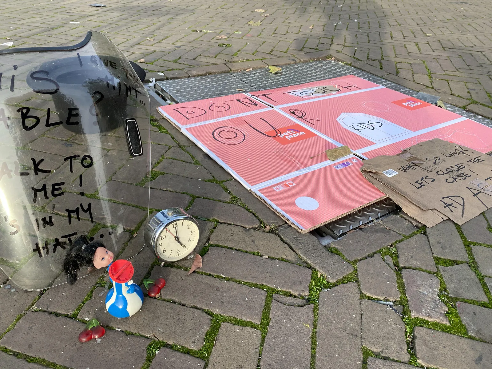 Without any context this is a rather strange scene on a street. There’s a piece of plastic with some unreadable words written on it. In front of this is a plastic doll and a small alarm clock that points at 5 o’clock. There’s also a piece of cardboard with a sentence written on it. It says Don’t touch our kids, why so long? Let’s close the case!