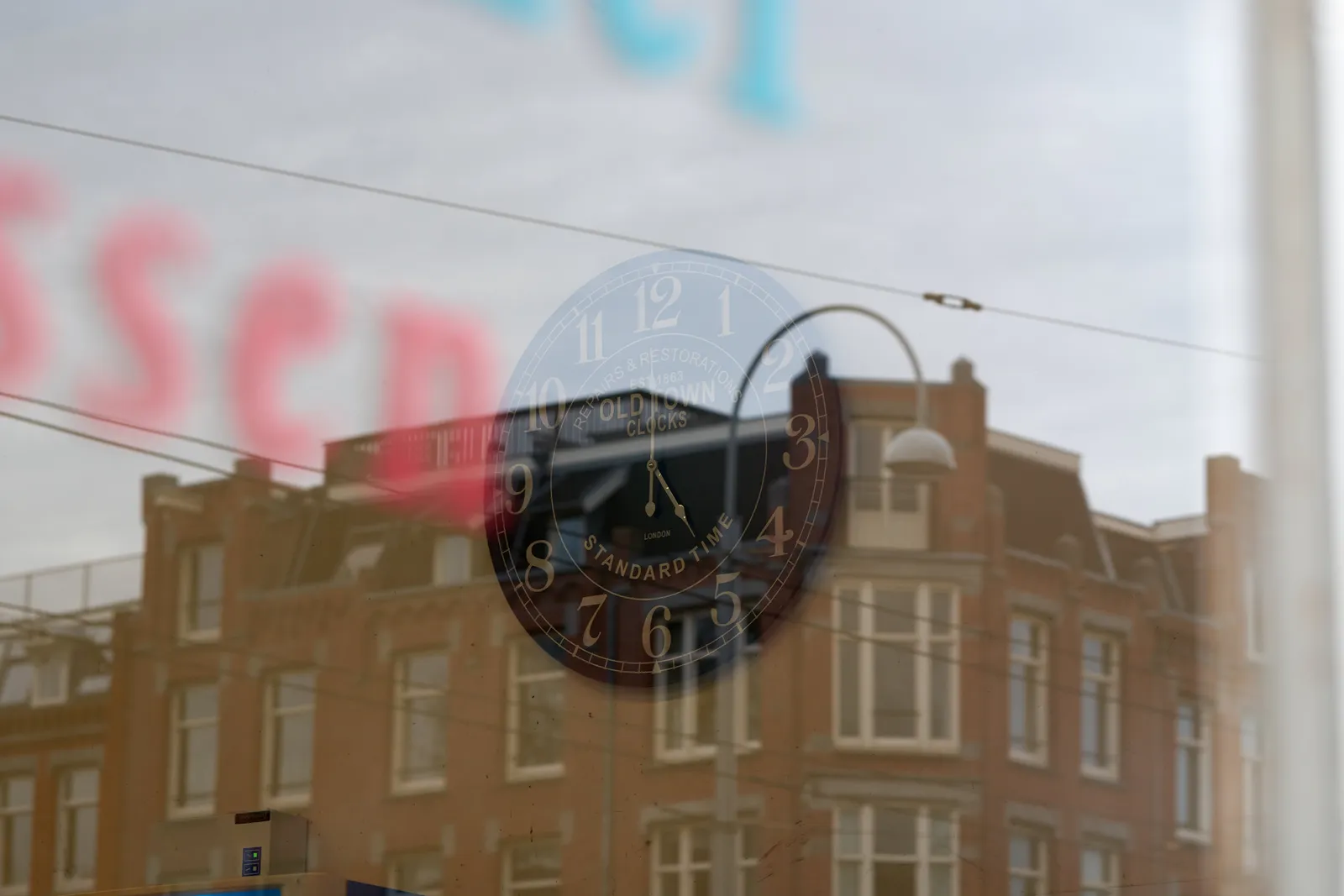 In the reflection of the shop window a building can be seen. On the window itself are some blurry letters. Through the window you can see a clock that points at 5 o’clock.