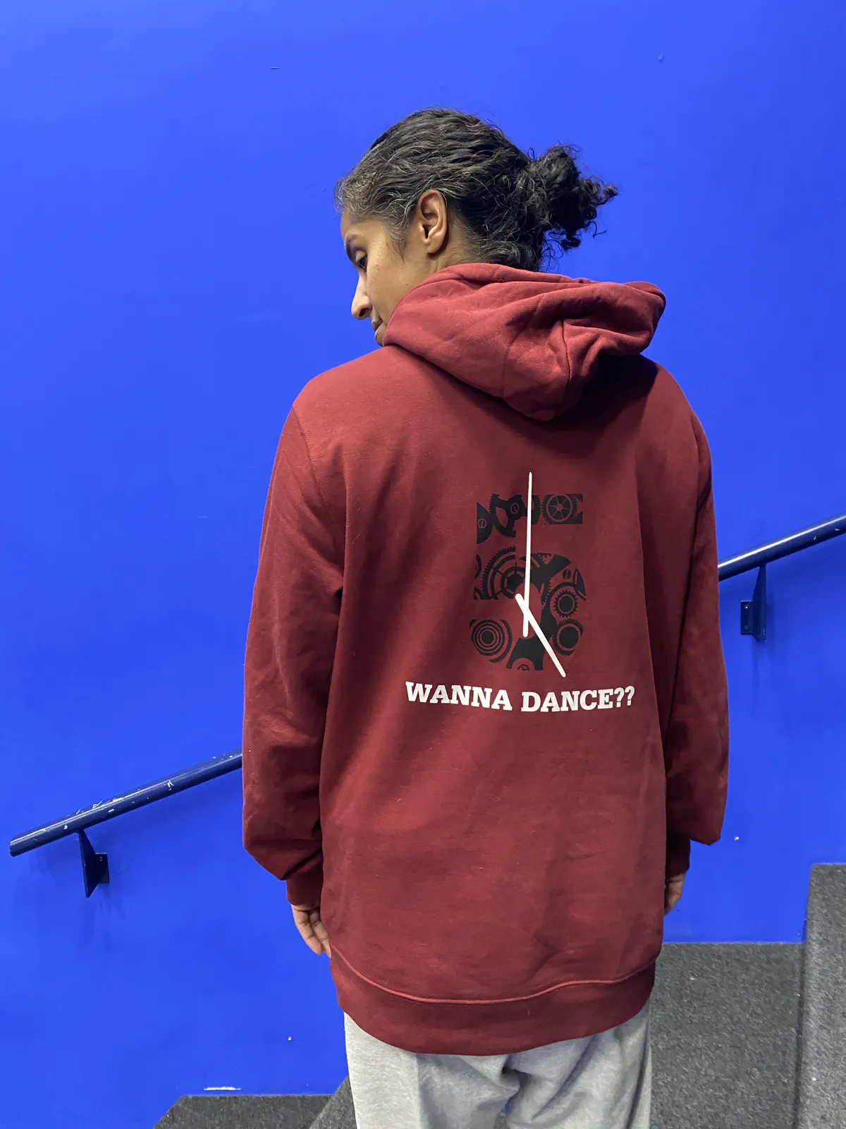 Looking at the back of someone in a red sweater standing in front of a blue wall. On the back sweater there’s a print that asks Wanna Sance? Above this question there’s a large number 5 with the hands of a clock printed over it. This clock points at exactly five o’clock. 