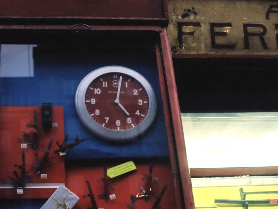 In a shop window, next to some swiss army knives, there’s a clock that points at two minutes past five.