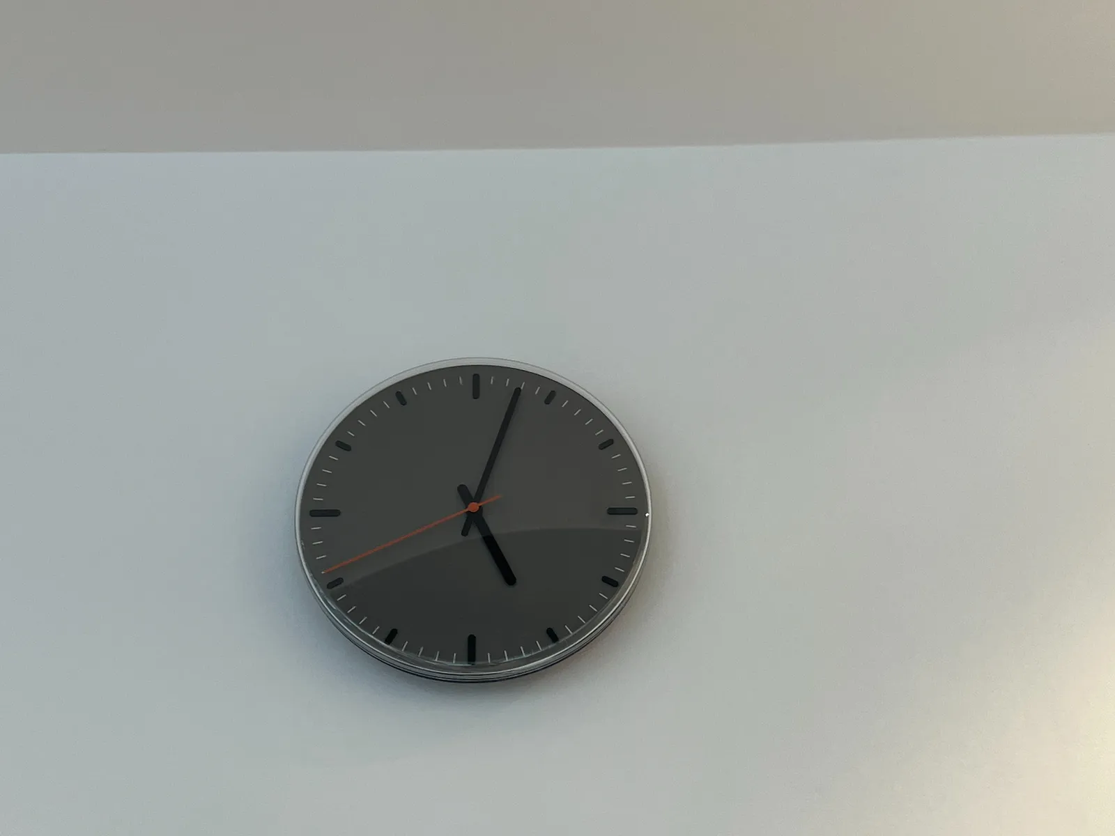 A minimalist grey clock on a grey and white wall. The clock points at 2 minutes past 5. 