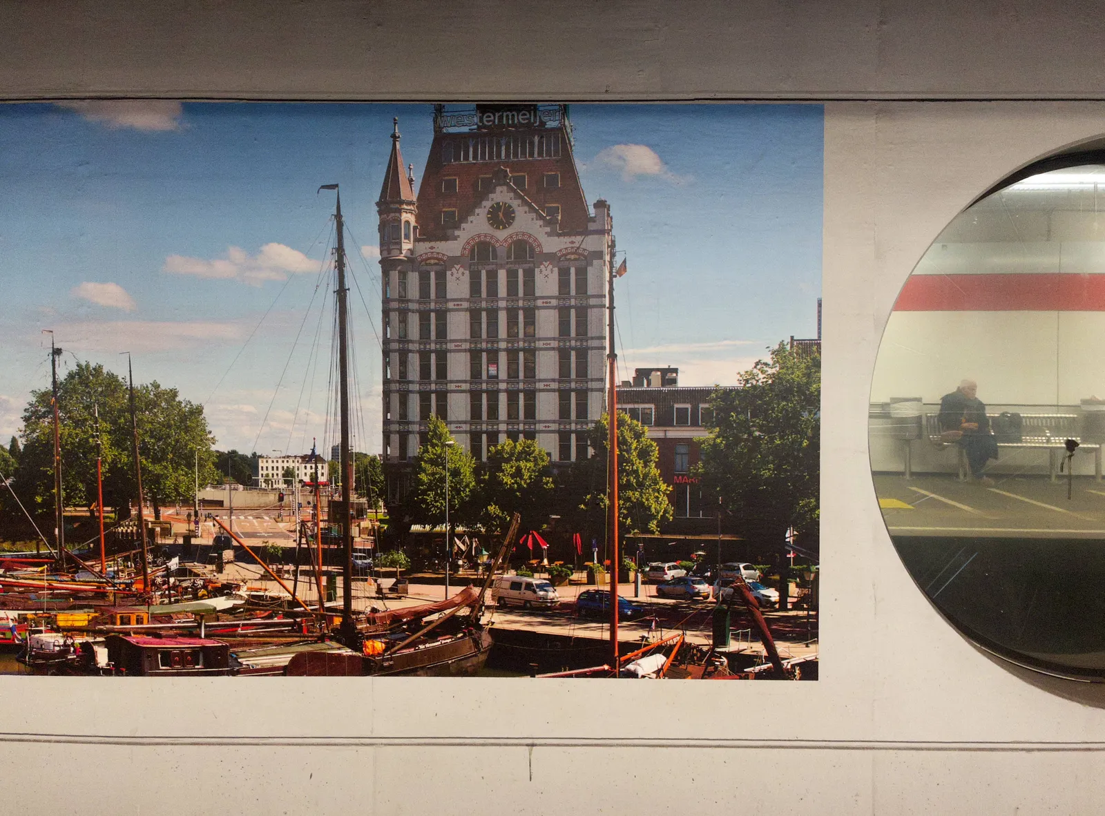 A picture of a wall, with on the right side a large round mirror, in which a man, sitting on a bench is reflected. On the left there’s an enormous photo of an older building with large wooden sailing yachts in front of it. There is a clock on this older building. It points at 2 minutes past 5.
