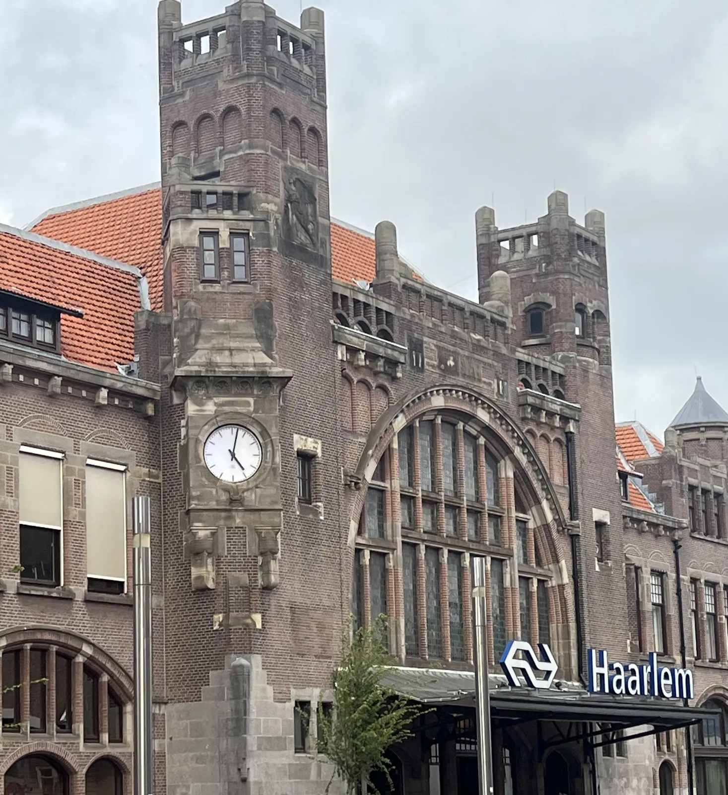 A large brick building which looks a bit like a castle, but according to the word and the logo on the facade is in fact the train station of the dutch city of Haarlem. There’s a clock built into this building and it points at 2 minutes past five.