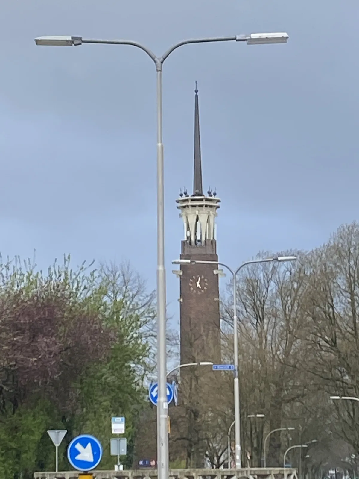 Looking past some street lights and some trees, in the distinace there’s a thin, high church tower with a clock on it that points at 2 minutes past 5.