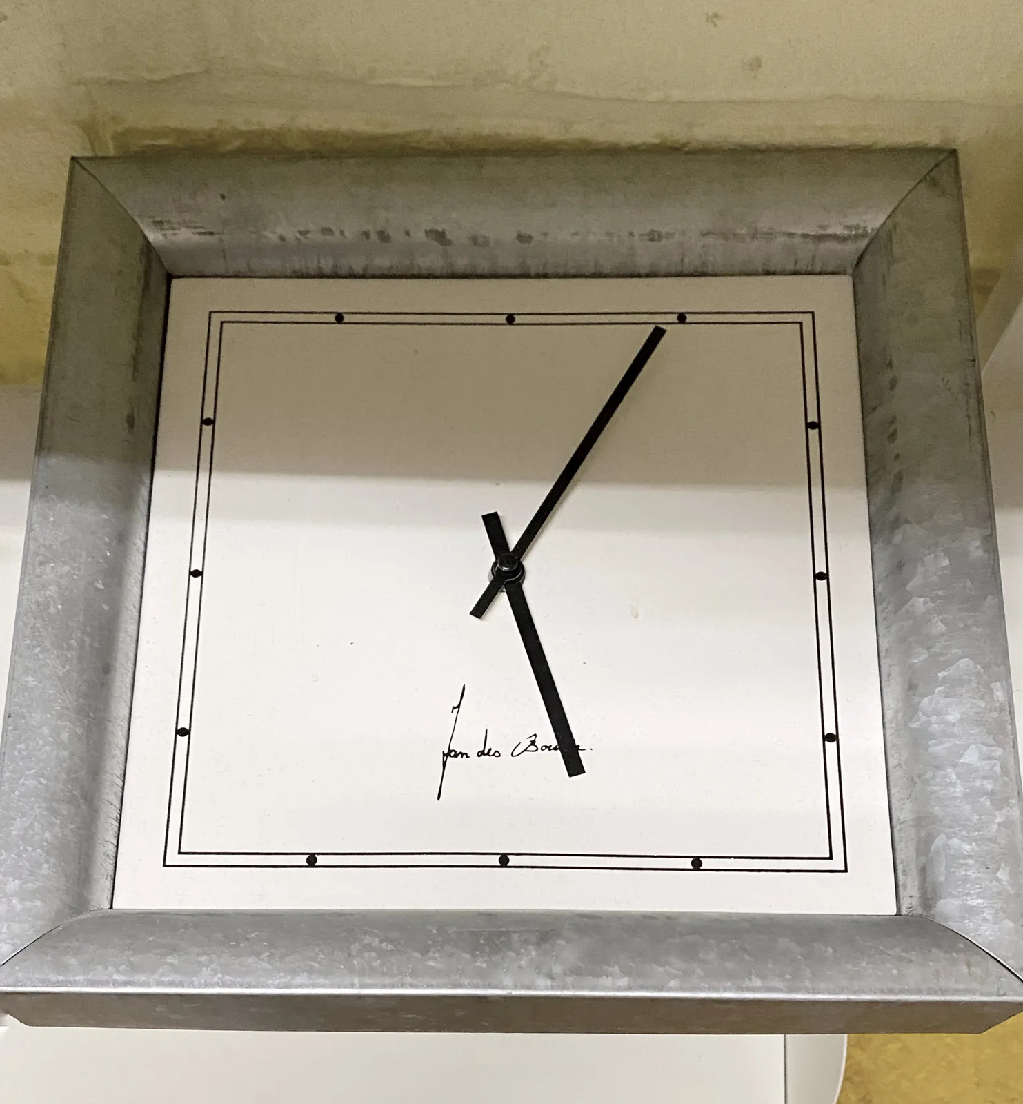 A white clock with black hands. It has an autograph printed on it by Jan des Bouvrie. It points at 4 minutes past 5.