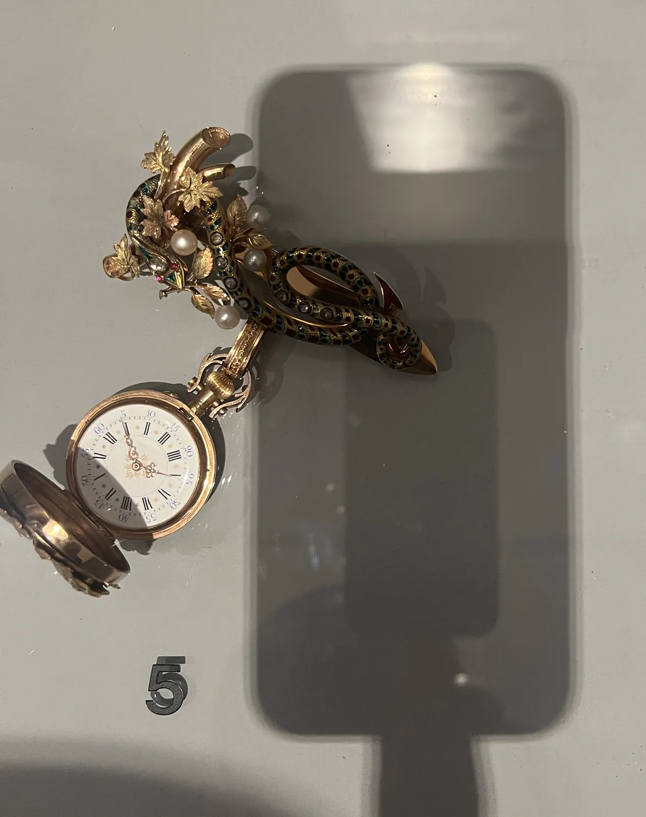 In a glass showcase there’s a richly decorated, golden, snake shaped brooch, with pearls and other precious stones, with a clock attached to it. The picture is taken with a phone, which is placed on the glass top, and the photographer is touching the phone with one finger. The shadow of this phone and finger take up the complete right side of the picture. The brooch with the clock is on the left. It takes a while to see what time it is, since the clock is not positioned straight in the picture frame. It points at 4 minutes past five.