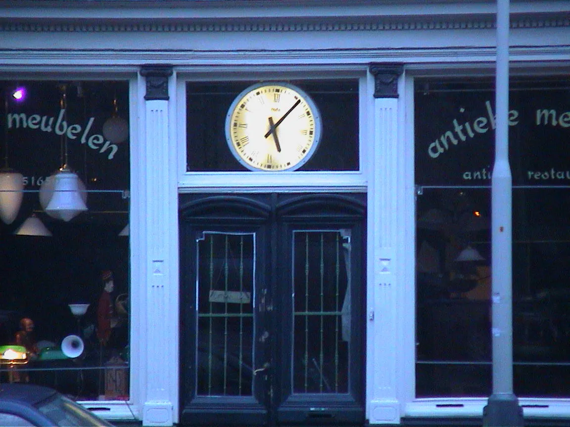 A large clock that’s a above the door to a shop that sells antiques points at six past five.