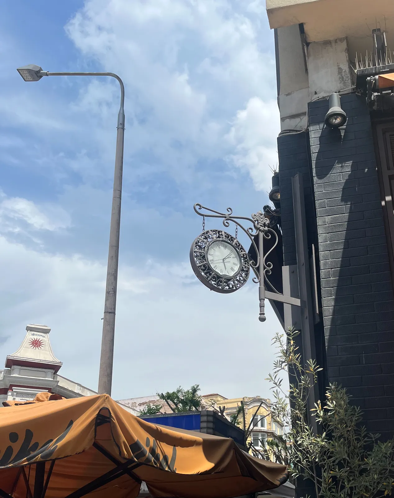 A clock attached to the corner of a building, on a sunny day. There’s a street light and a large parasol in this scene as well. The clock points at 6 minutes past five.