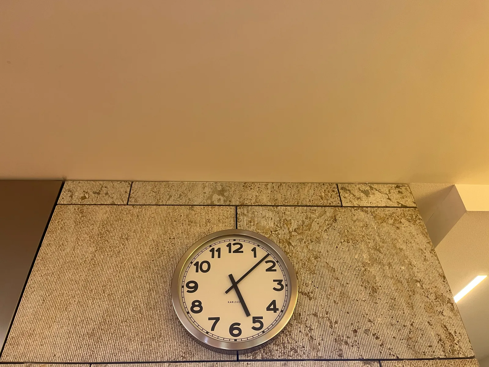 A large clock on a modern, gray stone wall. Part of the white ceiling can be seen as well. The clock points at 7 minutes past 5.