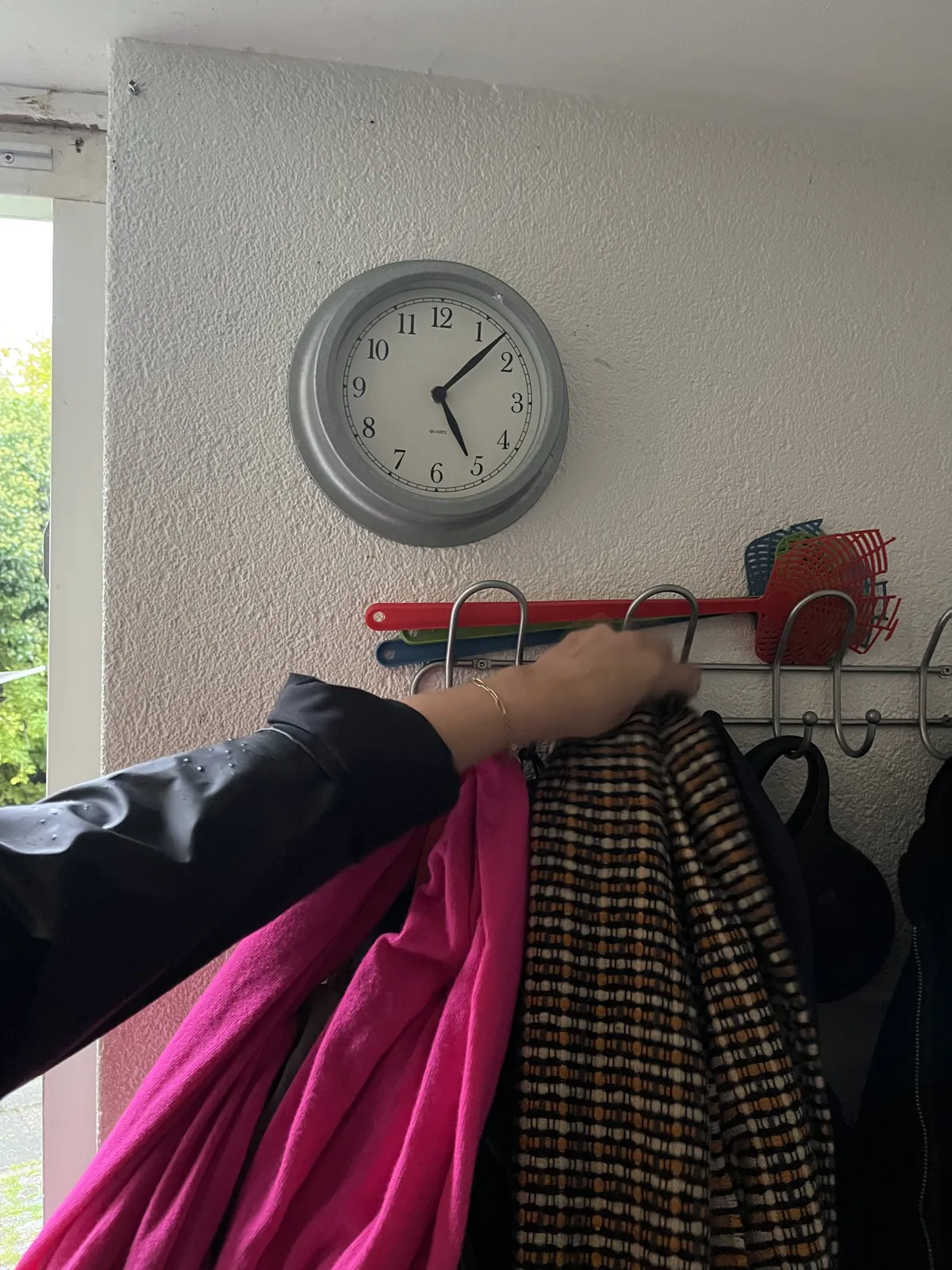 Someone is hanging their coat on a coat hanger. There’s a clock on the wall above the coats. It points at 7 minutes past five.