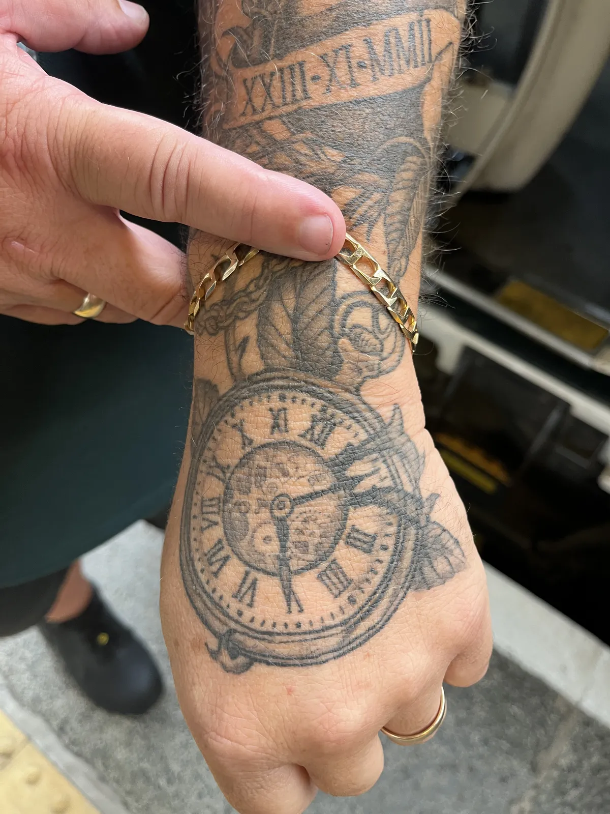 A tattoo of a clock on someone’s hand. There’s a date in roman numerals above it: XXIII - XI - MMII. The clock points at 8 minutes past five.
