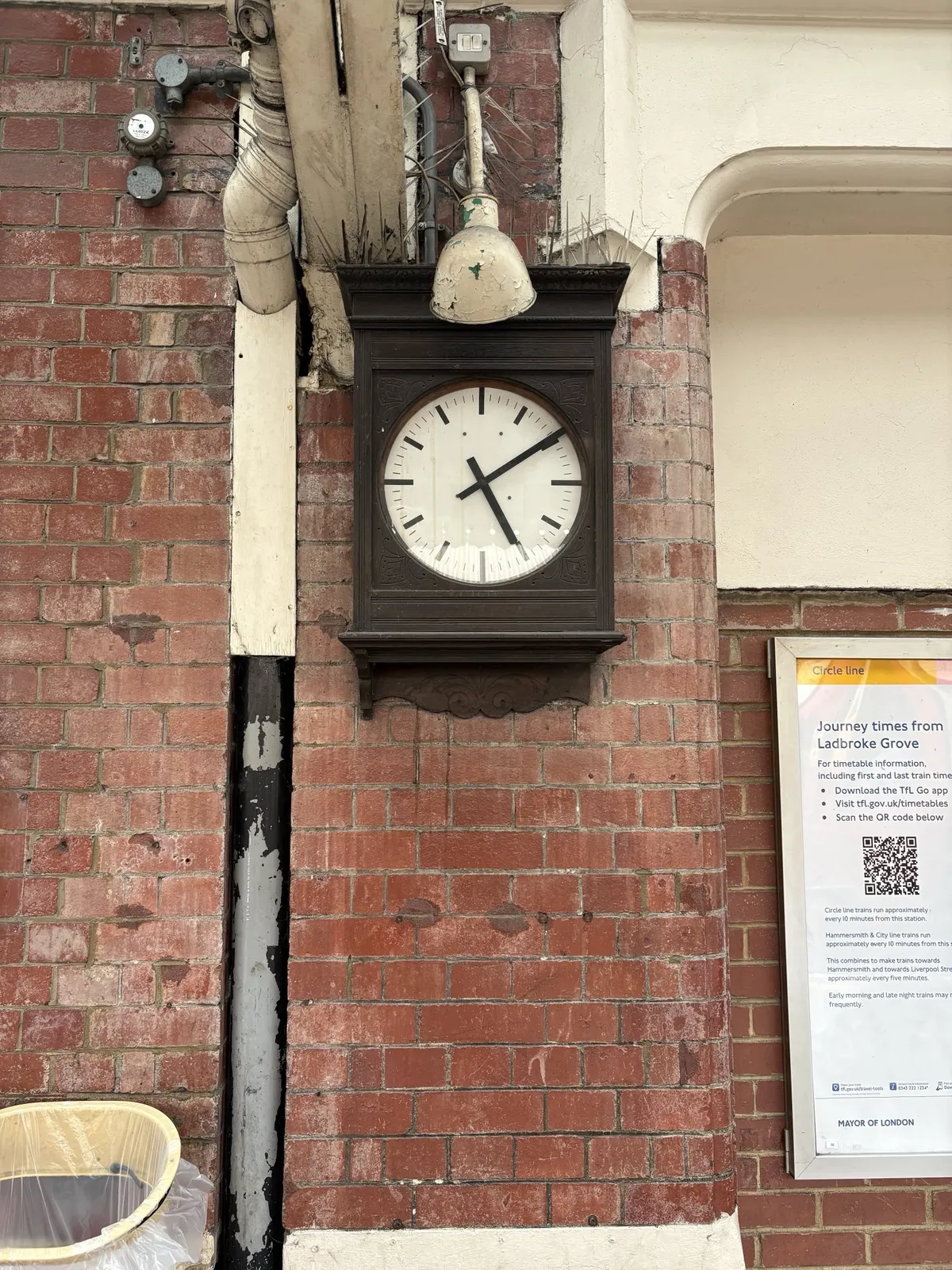 Drainage pipes, all kinds of wiring, and a clock on a brick wall. The clock points at 10 past five. 