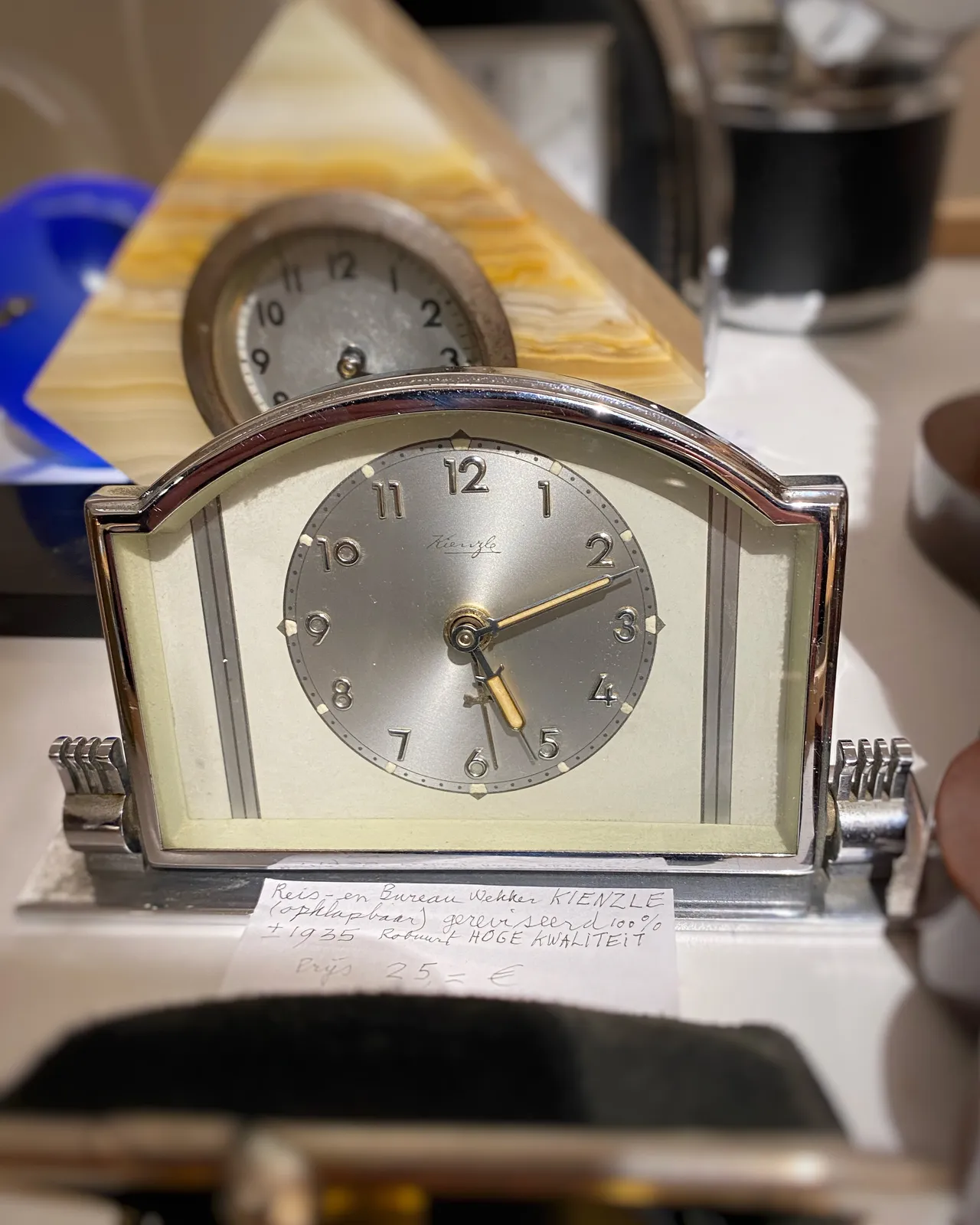 An old silver travel clock. It points at 11 minutes past 5. You can’t see the time on the clock that stands behind it. 