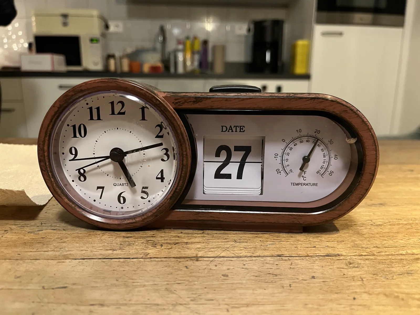 A device which shows the date in flipping numbers in the middle (27), the temperature with a dial on the right (22 degrees celcius), and the time on an analogue clock on the left. It points at 12 minutes past five.