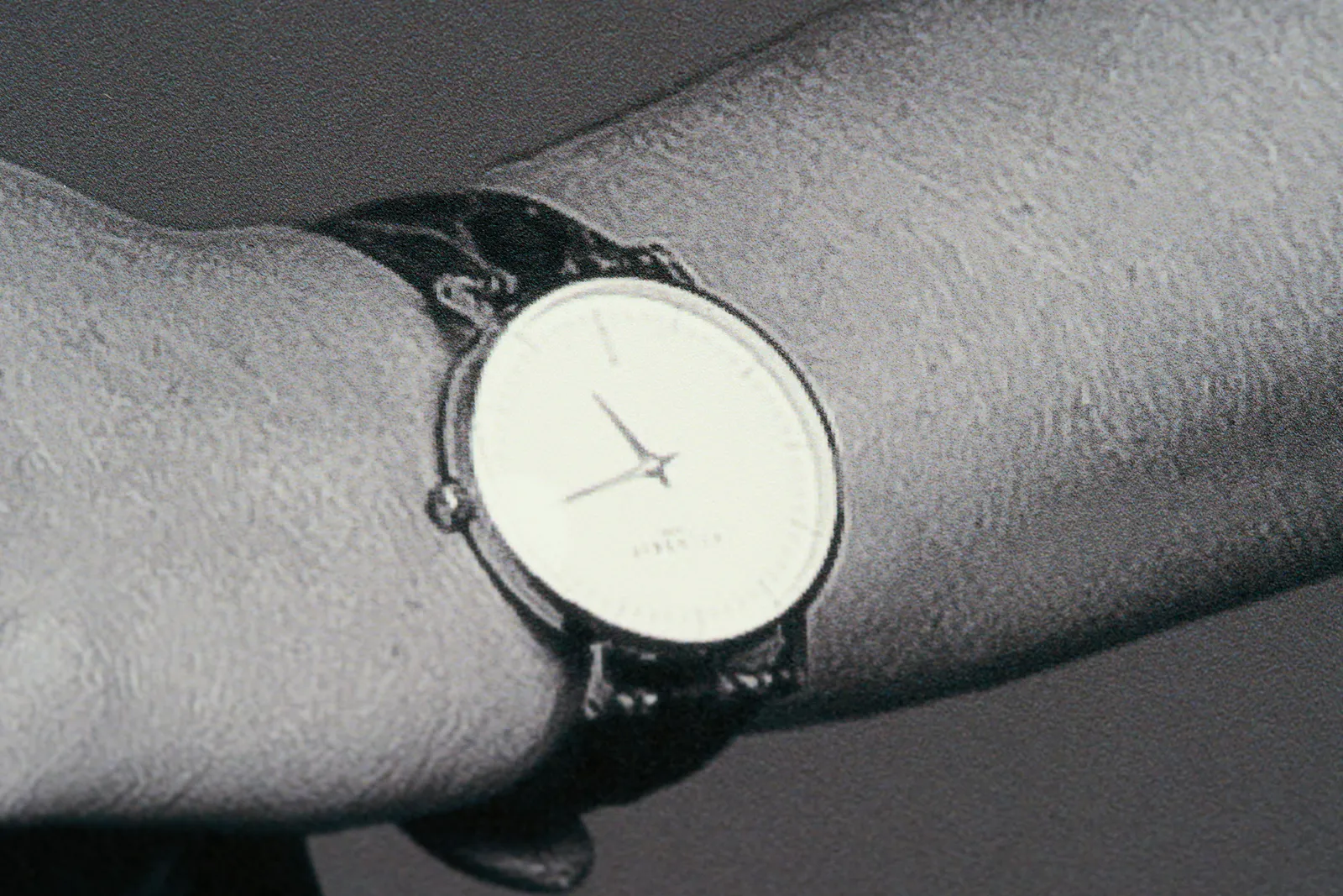 A picture of a detail of a picture. An arm with a watch. This watch is upside down so it takes a little time to see that it points at 13 minutes past 5.