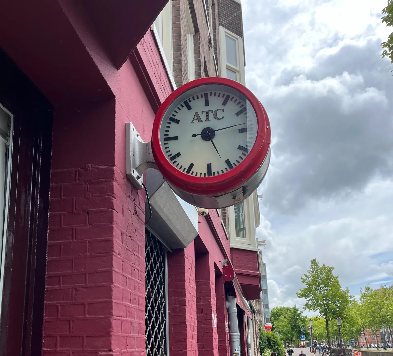 There’s a clock attached to this facade that’s painted red, in a street with trees. The frame of the clock is red as well, and it has the logo of a company called ATC on its face. There are three crosses, or exes, below each other inside the letter T, which may very well indicate that this is an Amsterdam based company. The clock points at 13 minutes past five.