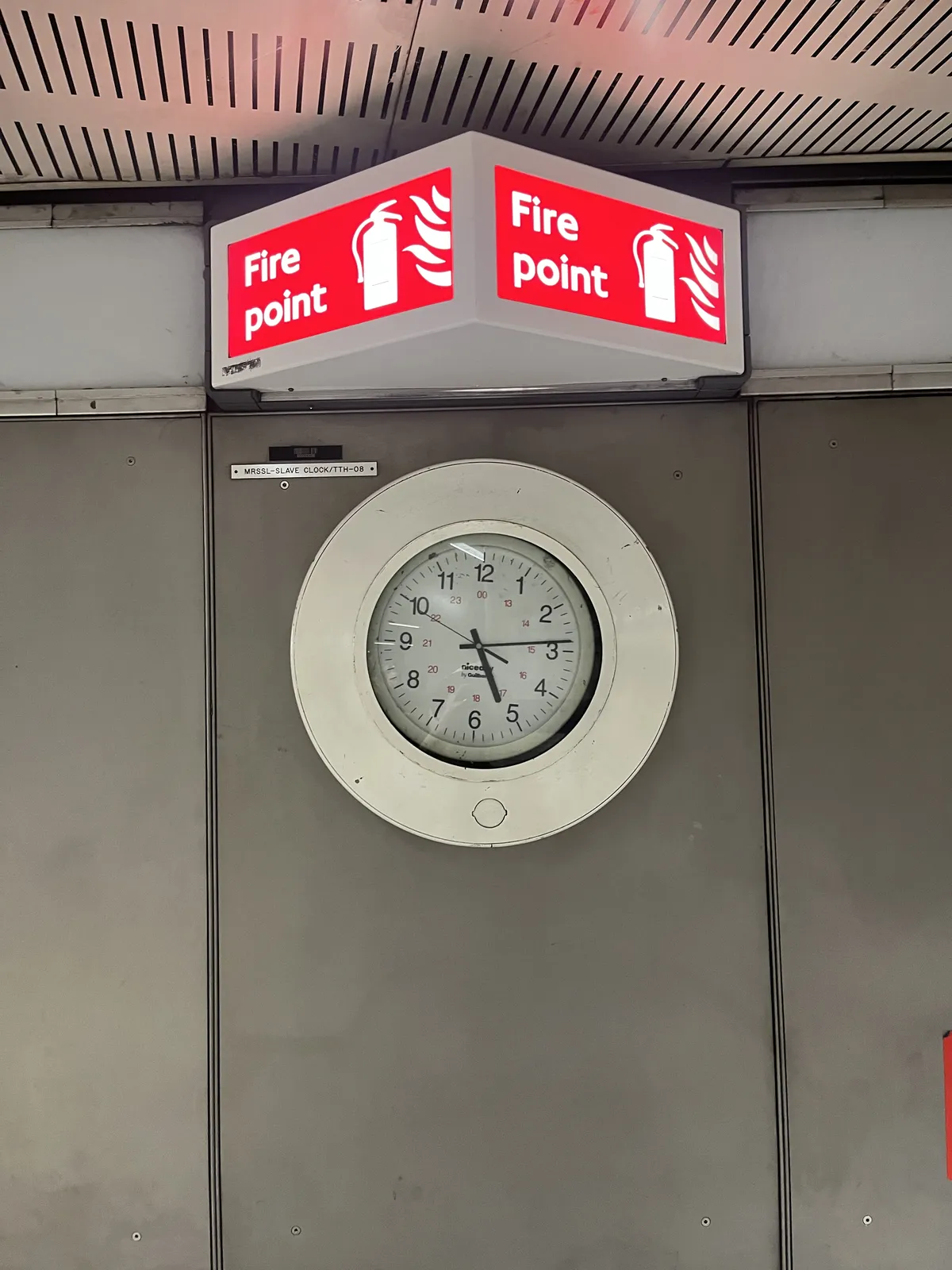 A clock, a bit off center in a round frame. Above it is a bright red sign that says Fire Point in white letters, with an icon of a fire extinguisher next to it, and some flames. The clock points at 14 minutes past five.