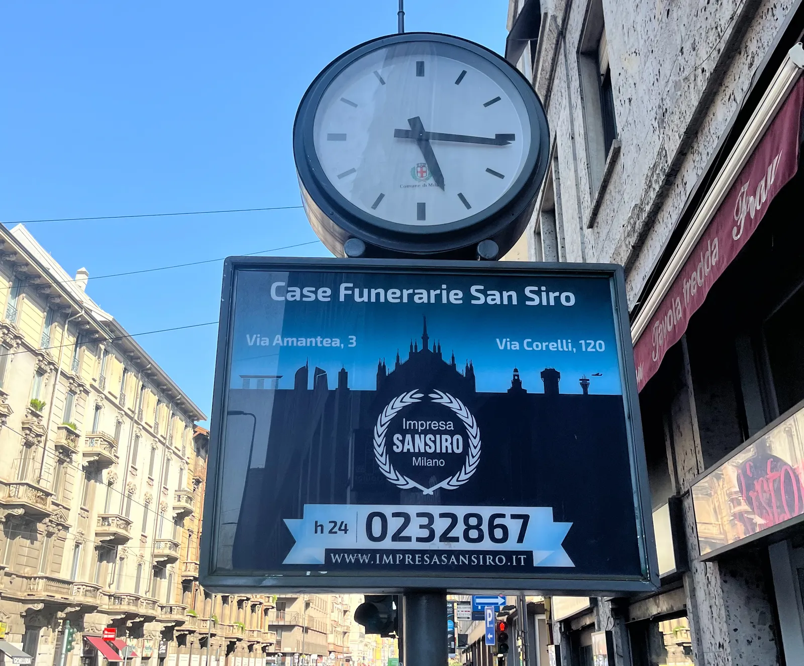 A clock on a pole, above an advertisement for Casa Funerarie San Siro in Milano. The clock points at 16 minutes past 5.