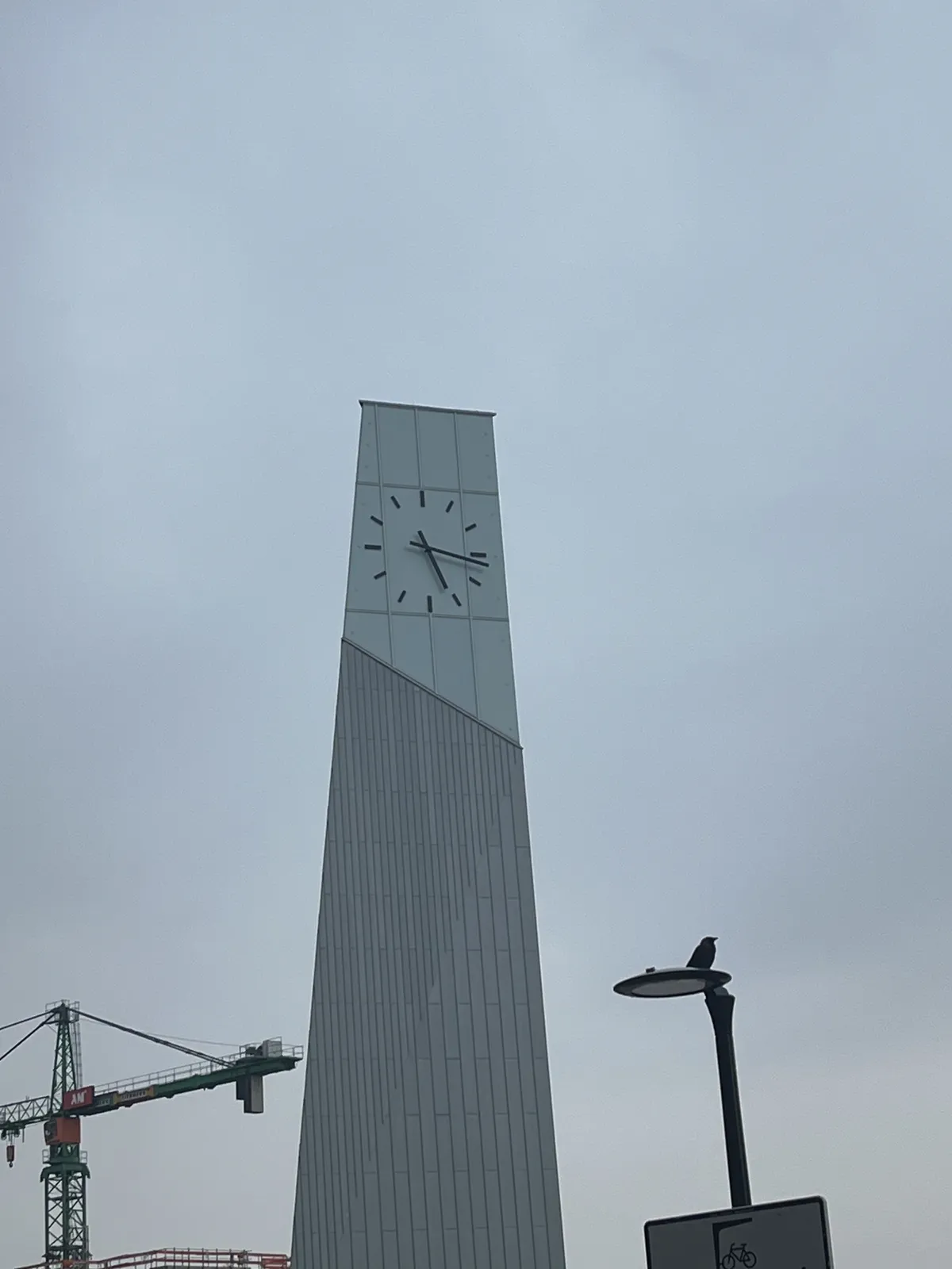 Against a gray, cloudy sky there’s a high crane and a lamppost. There’s also, in the middle of the image an enormous, modern tower with a modern clock on it. It points at 17 minutes past 5.