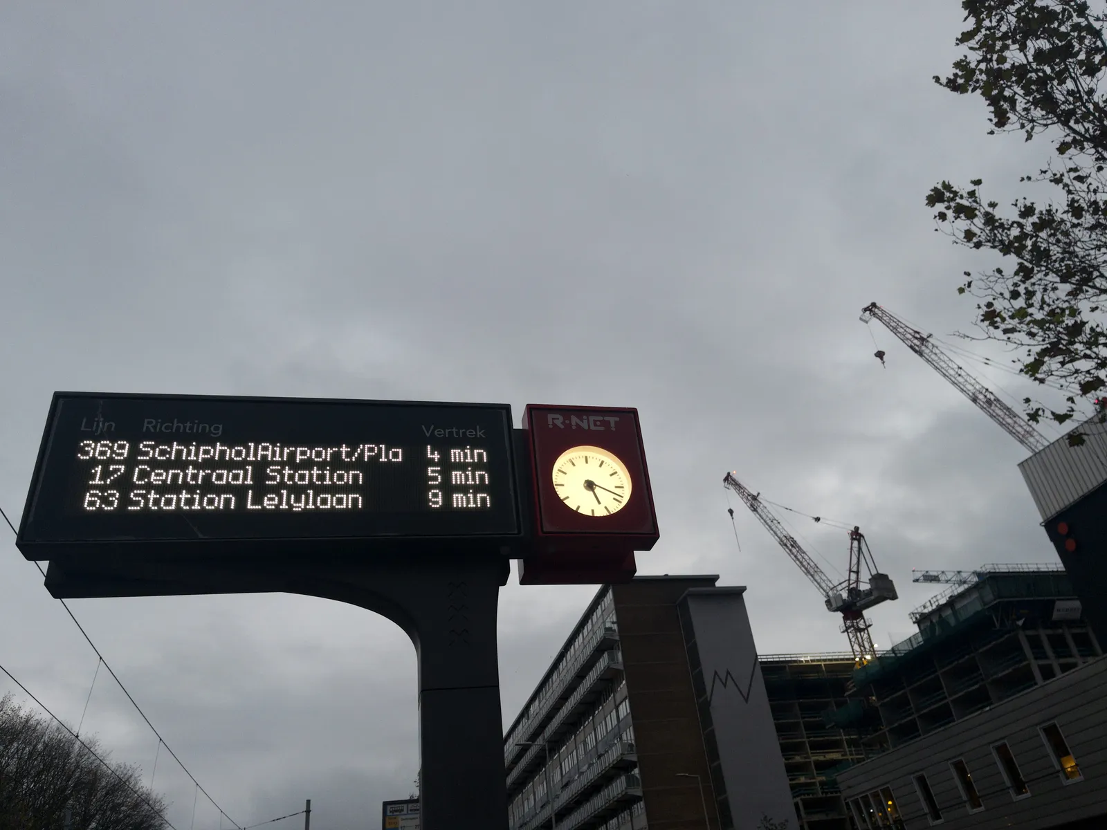 In the background is a very large appartment building with two huge cranes behind it. In the foreground a display that says that bus 369 to Schiphol Airport leaves in 4 minutes. The bus to Central Station leaves in 5 minutes and the bus to station Lelylaan departs in 9. The little clock next to this display shows that it is 18 minutes past 5.